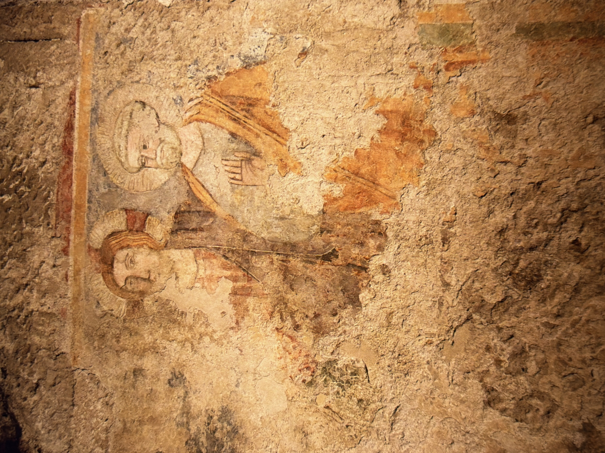 This is a close-up, eye-level, medium shot of a fresco depicting two figures on a textured, weathered wall. The fresco is faded and damaged, with areas of missing paint and visible cracks and imperfections in the plaster.
The figure on the left has a round face, reddish-brown hair, and a halo. They are depicted from the chest up, wearing a garment with some red and pink coloration. The figure on the right is also shown from the chest up, wearing a tan garment and a halo. This figure has a beard, a light-colored cap or head covering, and is shown with their right hand raised in a gesture of blessing or communication, with fingers spread. The figures are rendered in a simplified, early Christian style.
The background of the fresco is the rough, uneven surface of the wall, which is primarily in shades of beige, brown, and ochre. There are areas of darker discoloration and texture that suggest age and decay. The overall impression is of an ancient artwork in a state of considerable deterioration. This fresco is located in the catacombs of the Kyiv Pechersk Lavra monastery in Kyiv, Ukraine.