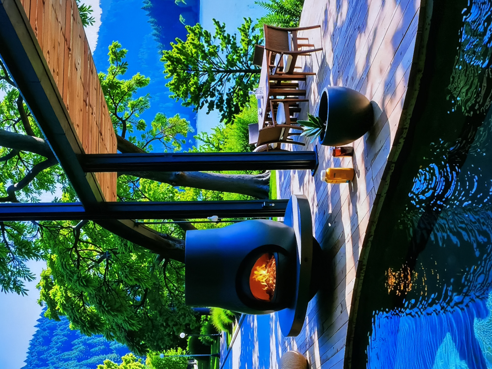 An inviting outdoor living space, located in Sofiivska Borshchahivka, Ukraine, features a wooden deck bordering a tranquil body of water, likely a pool or pond. The deck is furnished with a modern, dark-colored outdoor fireplace, emitting a warm orange glow from its burning fire, and a dining set comprising a table and several chairs. Two glasses of amber-colored beverages, possibly beer, are placed on the deck near the fireplace. A large, dark planter containing a succulent plant adds a touch of greenery to the deck area. Lush green trees and vegetation prominently surround the space, creating a vibrant natural backdrop, with distant hills or mountains visible under a bright, clear blue sky. The water reflects the sky and surroundings, with distinct ripples. The scene is bathed in bright daylight, suggesting a sunny day, and evokes a sense of relaxation and comfort. No text is visible in the image.