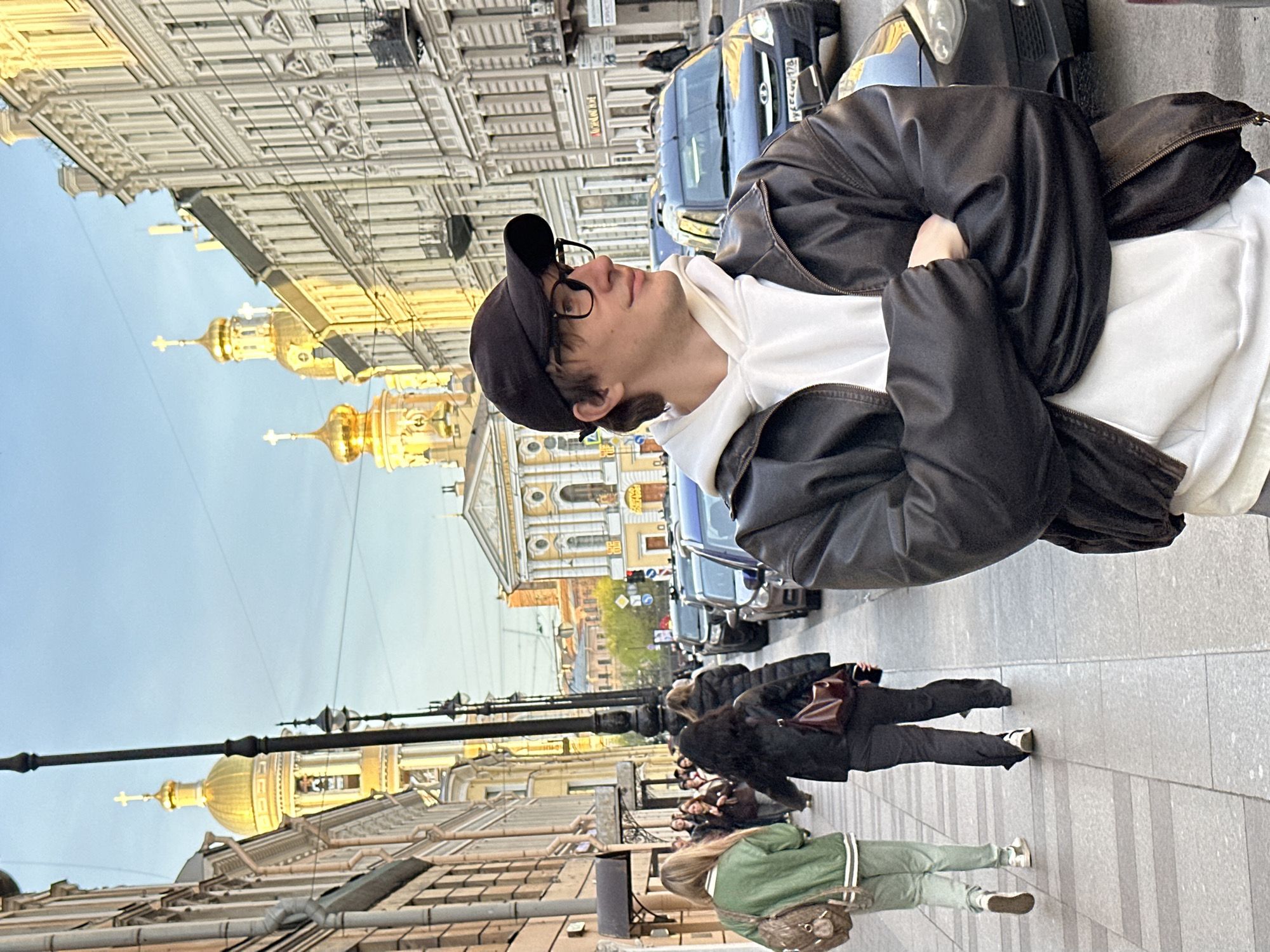 The image depicts a vibrant street scene in Saint Petersburg, Russia, during what appears to be late afternoon or early evening on a clear, sunny day. The primary subject is a young man in the foreground, seen from the waist up. He wears a black baseball cap, clear-rimmed glasses, a white hoodie, and a black leather jacket, with his arms crossed. He is looking upwards with a calm, contemplative expression, seemingly observing the surroundings.
The background showcases the distinctive architecture of Saint Petersburg, with multi-story historic buildings featuring intricate facades. Prominently visible against the clear blue sky are several golden domes, each topped with an Orthodox cross, characteristic of Russian churches or cathedrals.
On the street, pedestrians are visible walking along the sidewalk, while a row of parked cars lines the curb to the right. A blue van's license plate, "A004EC 178," is clearly readable, with "178" confirming the location as Saint Petersburg. Street lights, utility poles, and overhead wires are also part of the urban landscape. The scene captures a moment of daily life within the city's historic setting.