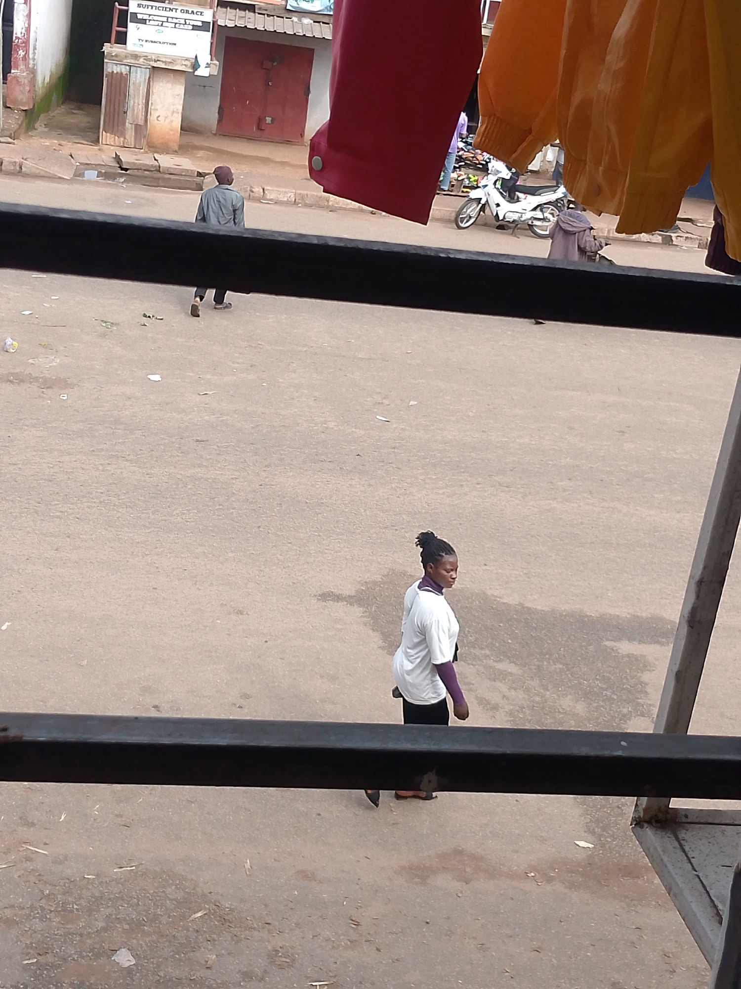 The image depicts a street scene in Bukuru, Nigeria. In the foreground, a dark metal bar partially obstructs the view. Beyond this, a woman with her hair in a bun is walking away from the camera, wearing a white t-shirt and dark pants. Further in the background, a person in a grey shirt and pants is also walking away from the camera. A white motorcycle is parked on the street. Various buildings, some with red doors, line the street, and there are some scattered debris and puddles on the ground. The overall environment appears to be an outdoor market or street.
