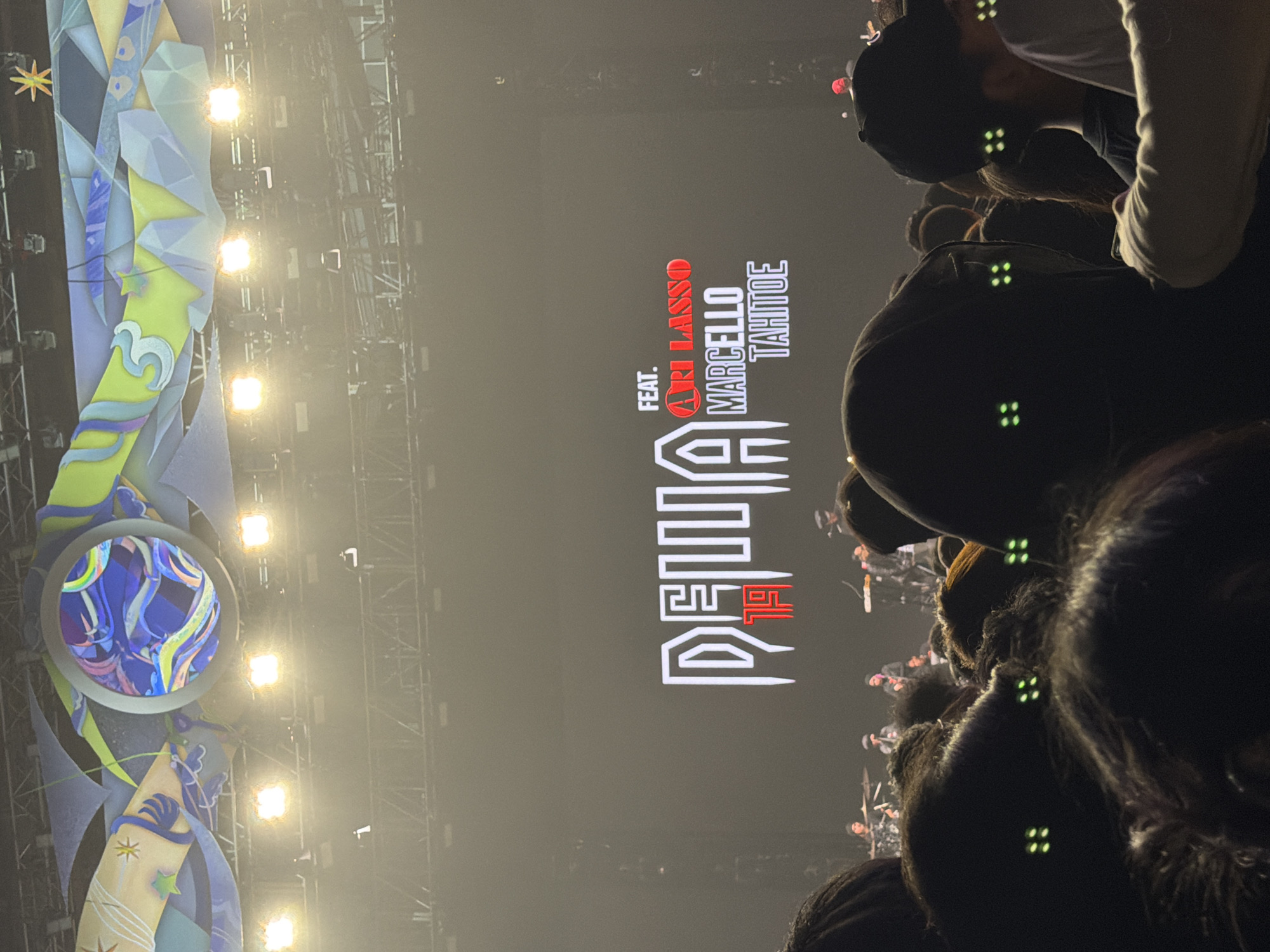 The image captures a concert crowd in Central Jakarta, Indonesia, with a large screen displaying the text "DELLA 19" in white, outlined lettering. Below this, "FEAT." is written in white, followed by "ARI LASSO" in red, and "MARCELLO TAHITOE" in white below that. The stage is lit with bright lights, and a band can be seen performing in the background. The foreground is filled with the heads of concertgoers, many of whom are wearing black and have green lights on their clothing or accessories. The overall atmosphere appears to be that of a lively music event.