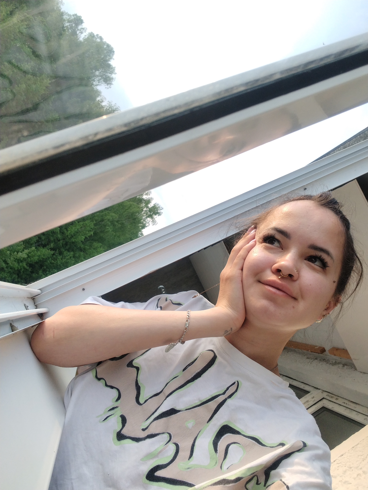 A young woman with dark hair pulled back, wearing a white t-shirt with a graphic design, is posing in front of a white, windowed structure. Her left hand is supporting her cheek. A bracelet is visible on her left wrist. Outside the windows, trees and sky can be seen. The location is not verifiable as Noviy, Russia without additional information.