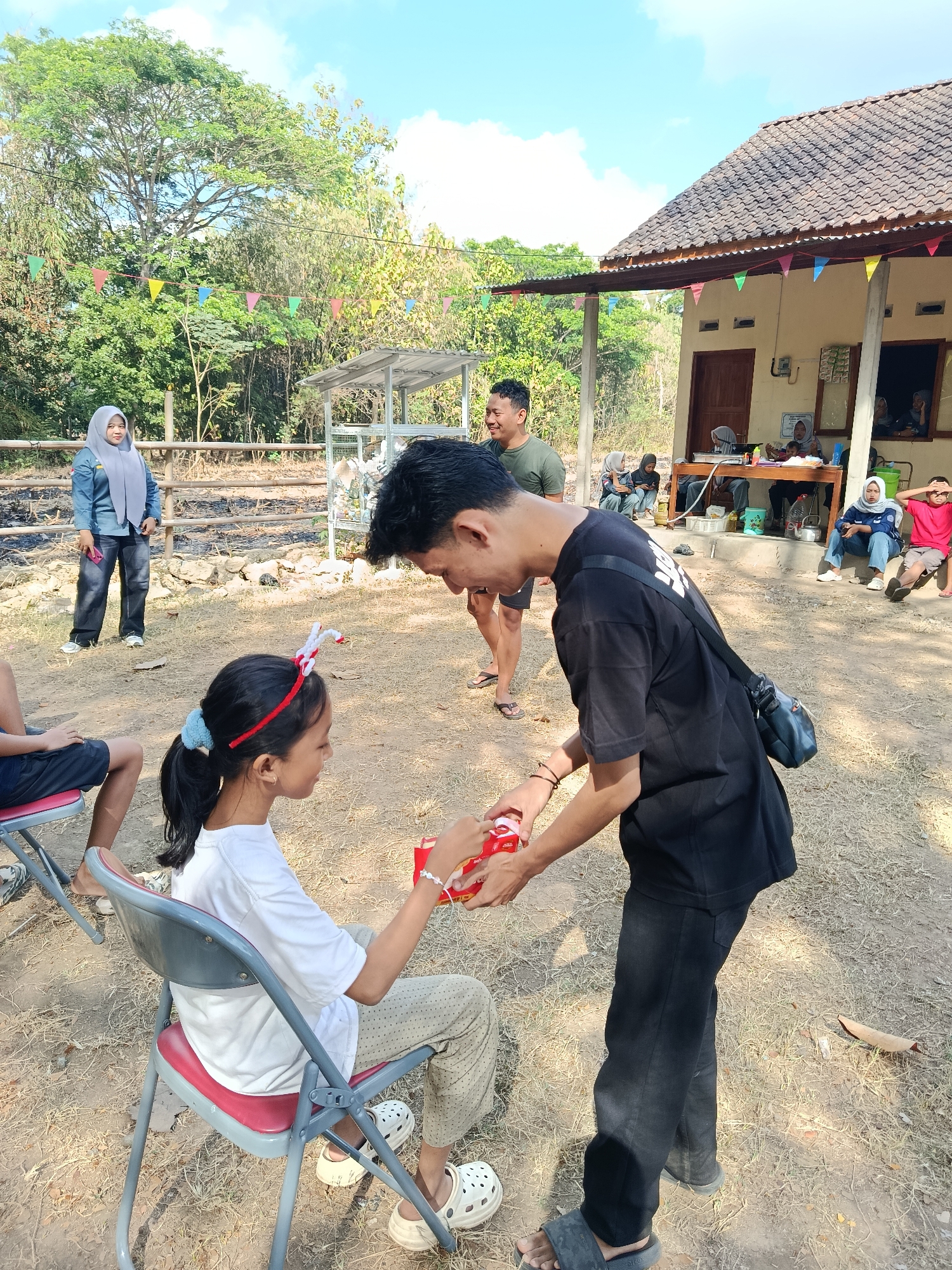 In Sedayu, Indonesia, a group of people gather outdoors in a rustic setting. A young girl, seated in a red and gray folding chair, is the focal point of the scene. She wears a white t-shirt and patterned pants, with a festive red and white headband adorning her dark hair, which is pulled back in a ponytail. Beside her, a young man in a black t-shirt and dark pants leans down, offering her a small red gift box.
In the background, a simple building with a tiled roof and an open window suggests a dwelling or community space. Several individuals are seated or standing around this structure, some wearing headscarves. Colorful pennant flags are strung across the scene, adding a festive touch to the dusty, sun-drenched ground covered in dry grass and scattered leaves. The environment is natural and somewhat unkempt, with trees and sparse vegetation in the distance, under a bright, partially cloudy sky. The overall impression is of a casual outdoor gathering or celebration.