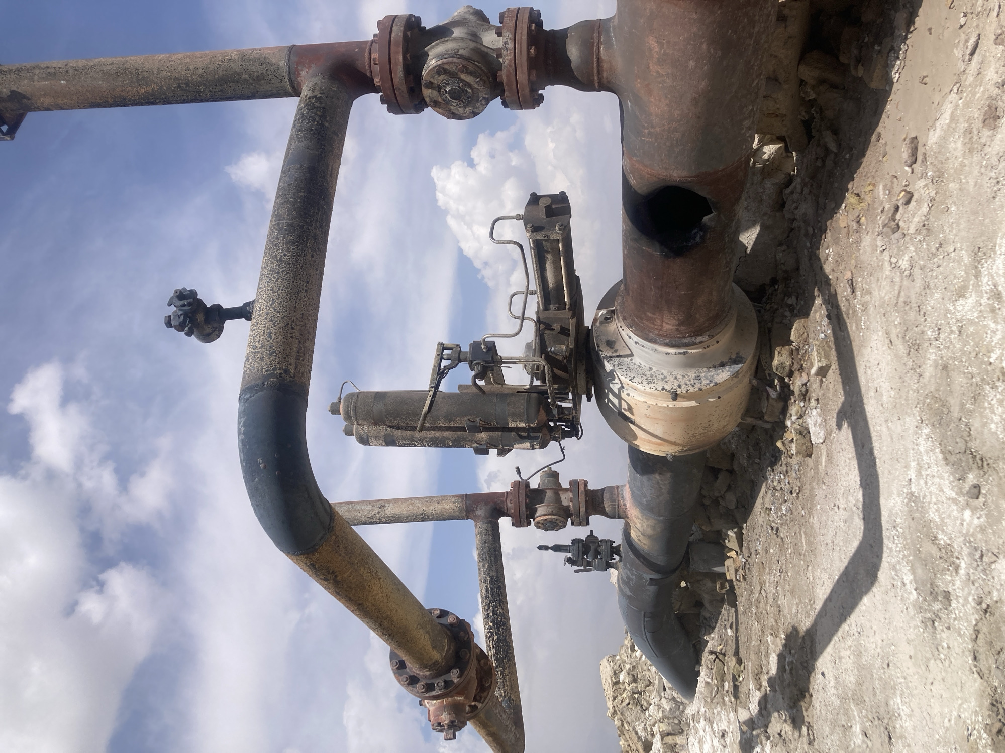 The image depicts a rusty, weathered industrial piping system against a background of a partly cloudy sky and a crumbling concrete structure. The pipes show signs of corrosion and aging, with patches of rust, peeling paint, and a significant hole in one section. Valves and other fittings are incorporated within the pipe network. The concrete structure appears dilapidated, with broken edges and a rough surface. The location in the city is Unknown, Unknown. There are no people visible in the image.