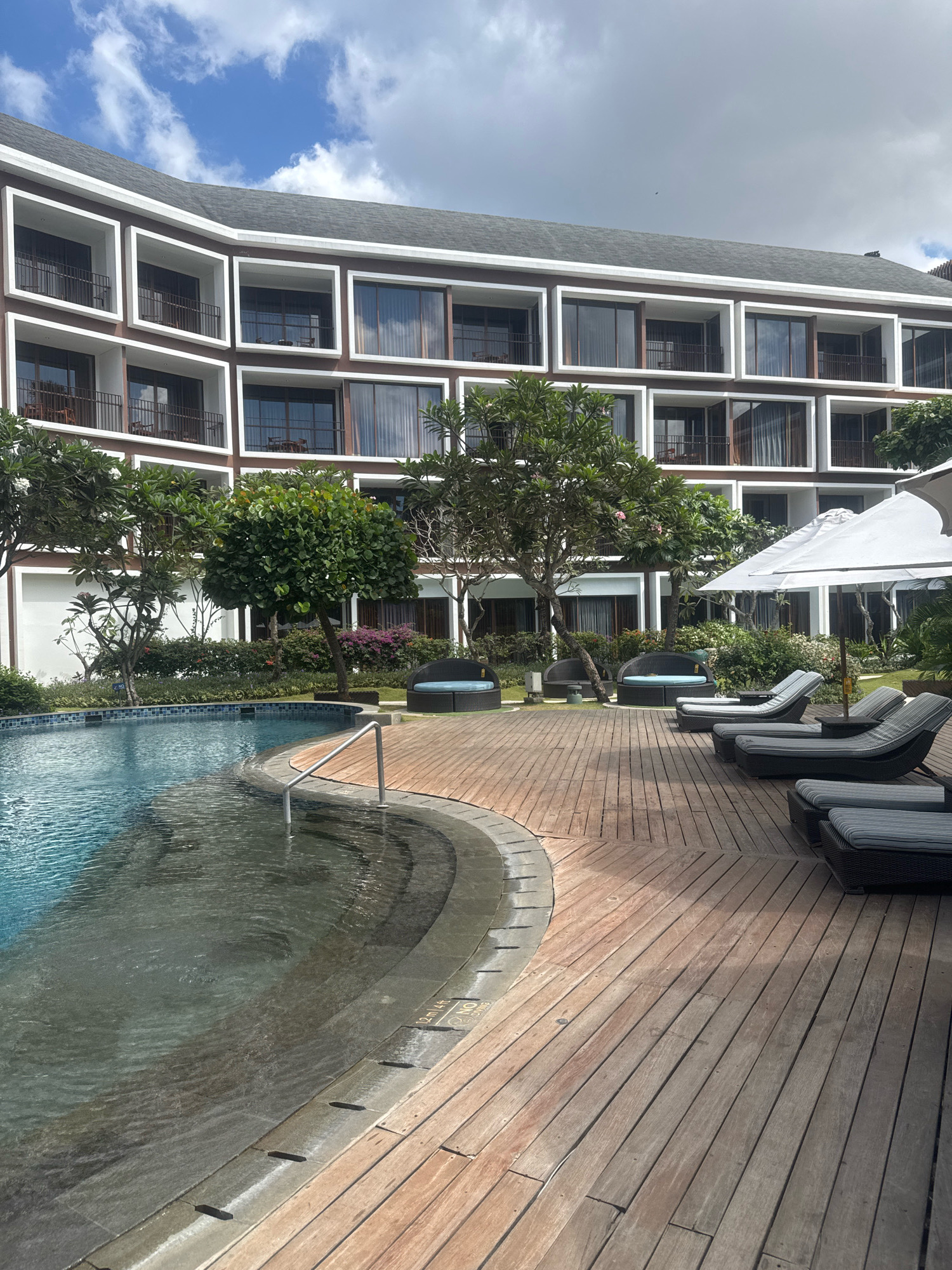 A wide-angle, eye-level outdoor shot captures a resort scene in Kuta, Indonesia. The primary focus is a curved swimming pool in the foreground, its water a vibrant blue. To the right, a wooden deck is furnished with several grey lounge chairs, some with striped cushions, and a large white umbrella. Beyond the pool, lush green trees and flowering bushes provide a natural border to the resort's architecture. The resort building itself is a multi-story structure with a distinctive modern design, featuring repeating rectangular window frames in a dark brown exterior. The sky above is partly cloudy with a mix of white and grey clouds. The overall impression is one of a serene and luxurious tropical getaway.