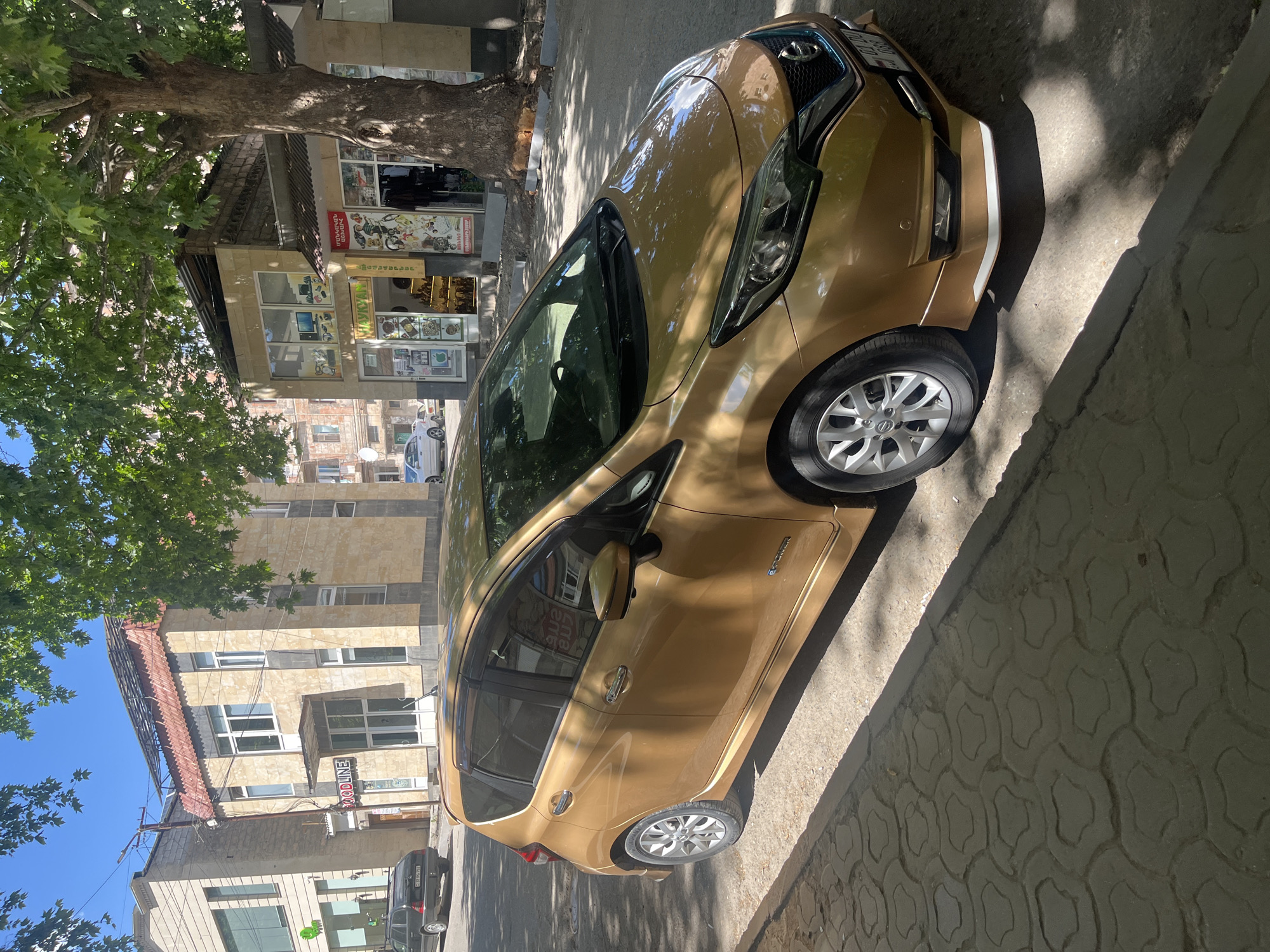 A gold Nissan Note is parked on a sidewalk next to a street in an urban environment. The car is facing to the right, with its front slightly angled towards the viewer. The sidewalk is paved with decorative grey interlocking pavers, casting shadows from a large tree. In the background, there are several buildings with storefronts. One building on the left has a sign that reads "FOODLINE". Another building to the right features a tree with a large trunk prominently in the foreground, and multiple storefronts with displays of merchandise. The sky is clear and blue.