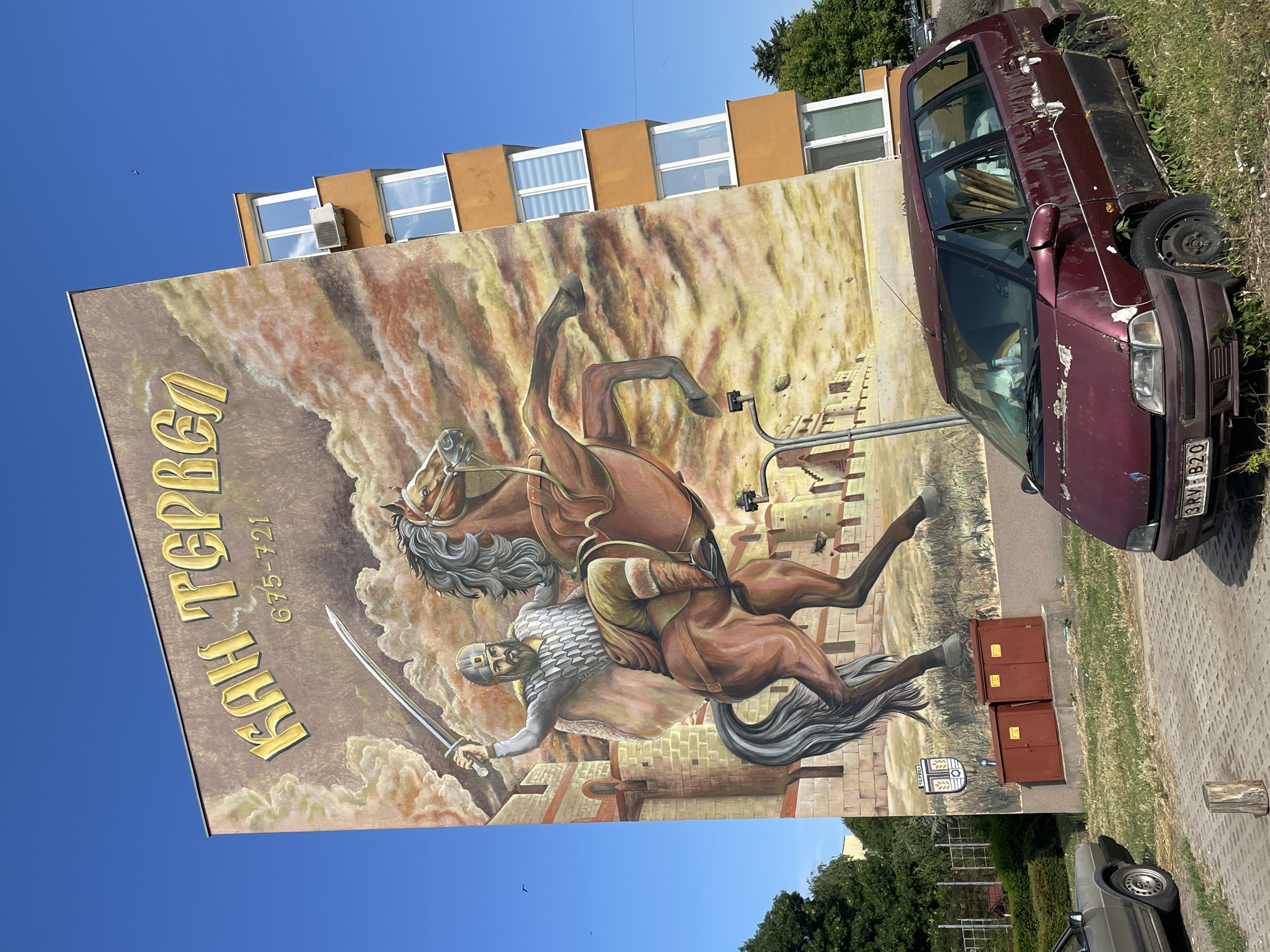 This is a photograph of a mural on the side of a building in the city of Tervel, Bulgaria. The mural depicts a historical figure, identified by the text above as "КАН ТЕРВЕЛ" (Khan Tervel), a Bulgarian ruler from 675-721. The figure is shown on horseback, raising a sword. The background of the mural features clouds and a castle. In the foreground, two red electrical boxes are visible. A dark red minivan is parked in front of the building, with its paint peeling and vegetation growing around it. To the left of the minivan, part of a light-colored car is visible. The scene is set on a paved area with grass and trees surrounding it. The sky is clear and blue.