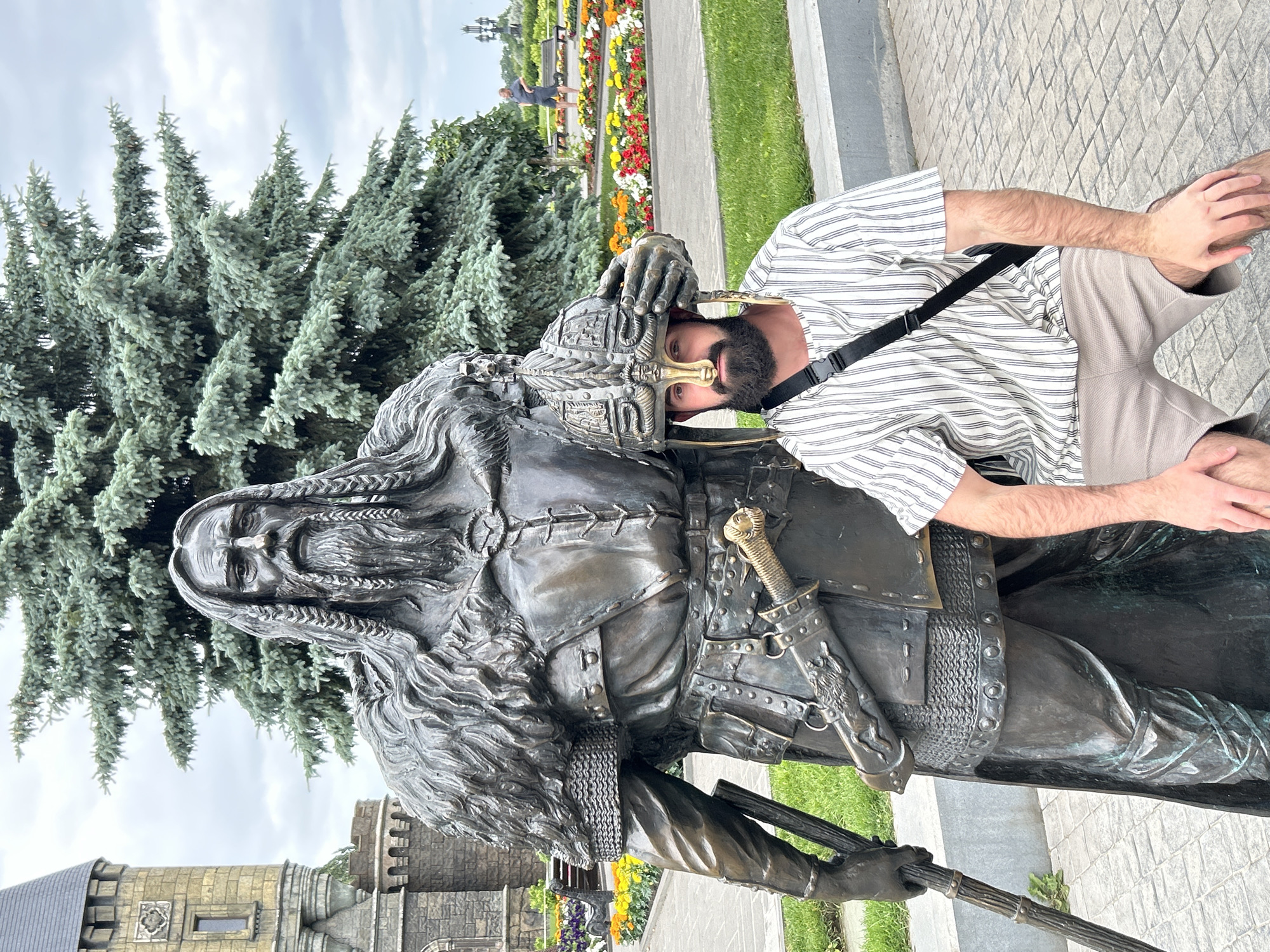 The image depicts a man posing with a bronze statue of a historical figure in Khryashchevka, Russia. The man is wearing a striped shirt and light-colored shorts. He is leaning against the statue, with his hand resting on its helmet. The statue appears to be that of a warrior, with a long beard, braids, and armor. It is holding a weapon. In the background, there are trees, a manicured garden with flowers, and a stone structure resembling a castle. The sky is overcast, suggesting it is daytime. The overall impression is that of a tourist visiting a historical or cultural site. There is no visible text in the image.