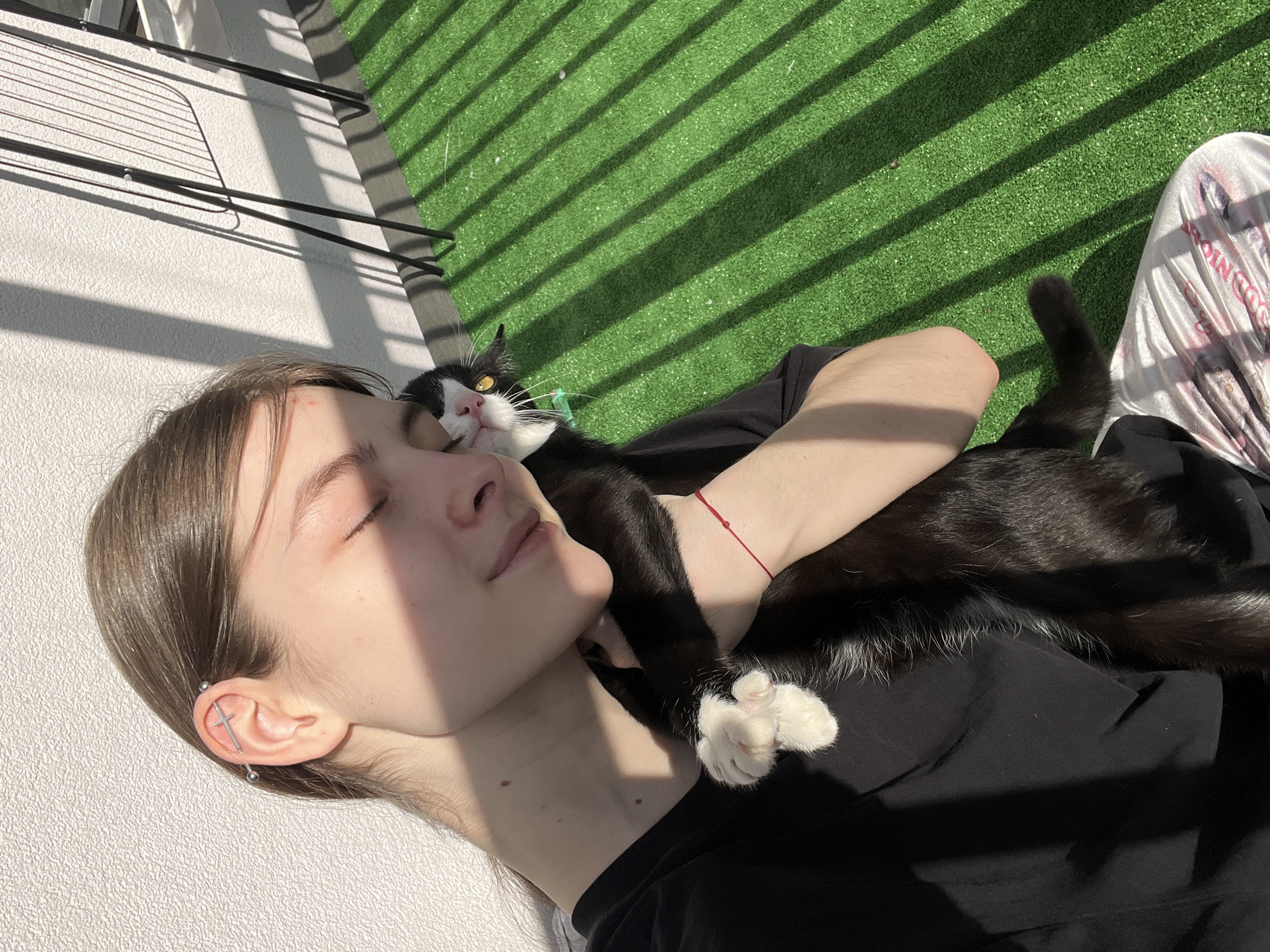 A young woman reclines with her eyes closed, face tilted toward the sun, on a balcony, with a black and white cat nestled on her chest. The woman wears a black shirt and has an industrial piercing in her left ear. A red string bracelet is visible on her left wrist. The cat has white paws and a green collar. The balcony floor is covered in artificial grass, and shadows from a railing cast stripes across the surface. A drying rack stands against the white wall on the left side of the image. The location in the city of Saint Petersburg, Russia, cannot be determined from the image.