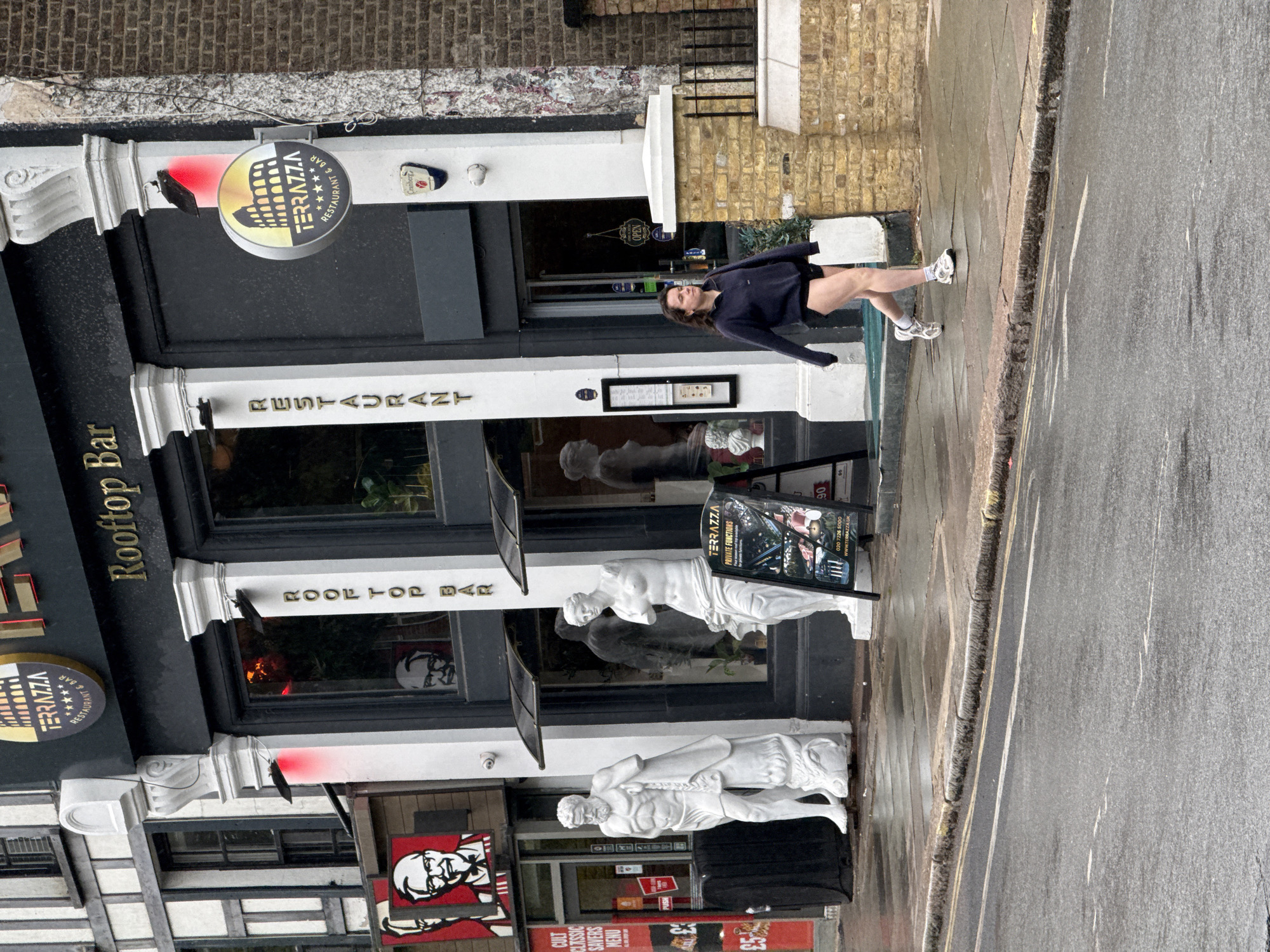 The image shows the exterior of the Terrazza Rooftop Bar and Restaurant in London, United Kingdom, with a woman walking on the sidewalk in front of the building. The restaurant has a black facade with white columns and gold lettering. There are statues of classical figures flanking the entrance. A KFC restaurant sign is visible in the lower left corner. The sidewalk and street appear wet. The woman is wearing a dark jacket and shorts.
