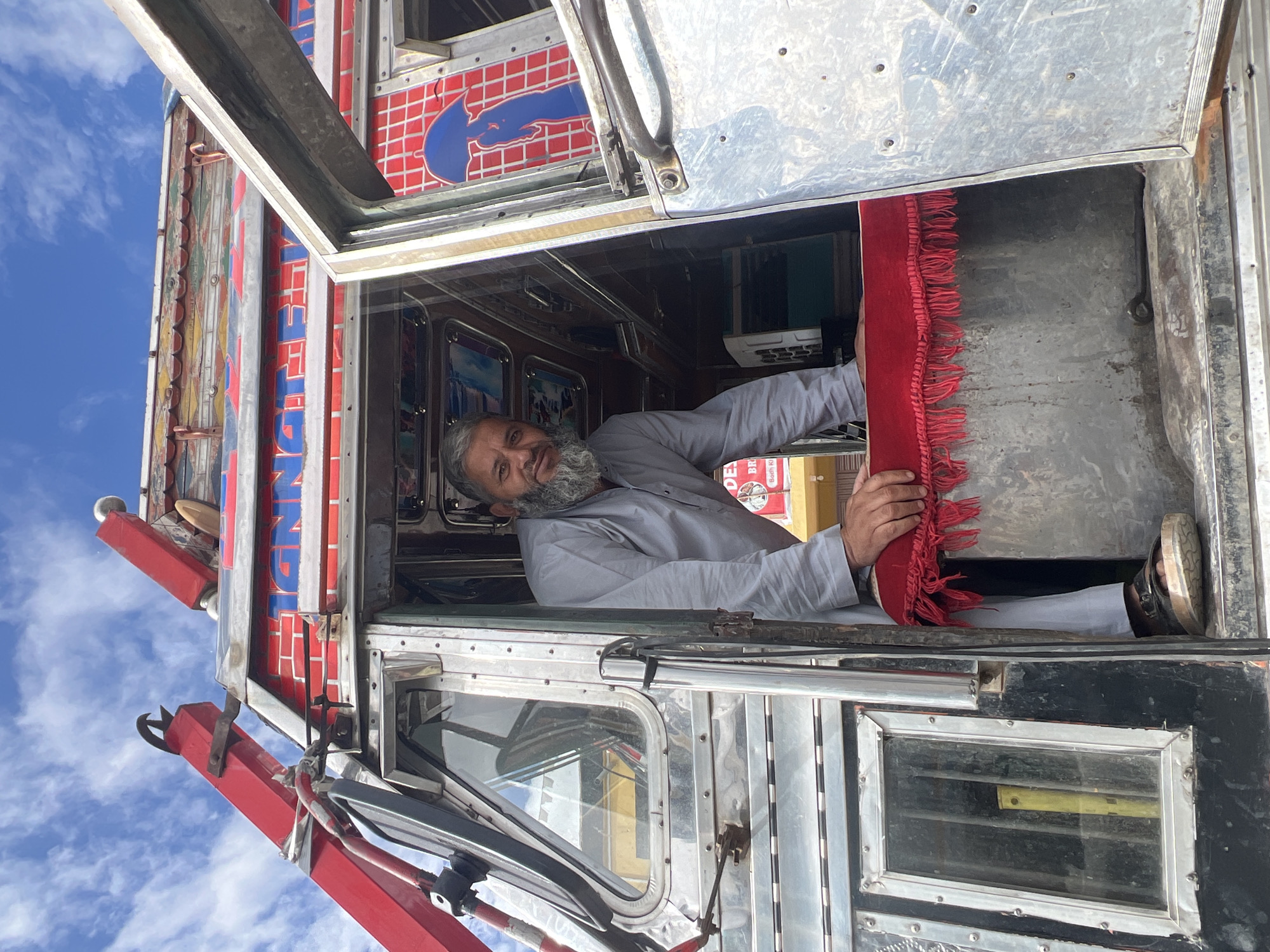 A man with a beard and gray shirt is standing in the open doorway of a truck in Bodhkharboo, India. The truck is decorated with red, blue, and white lettering and colorful designs. The man is leaning on a red fringed cloth that covers the top of a metal compartment. The truck has chrome accents and windows. The sky is blue with white clouds.