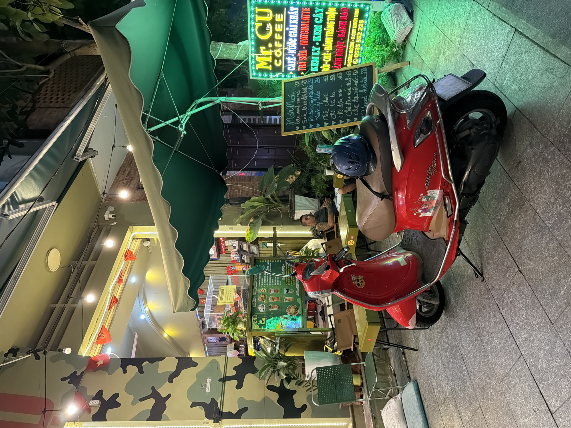 A red scooter is parked on a paved sidewalk in front of a cafe with camouflage-painted columns and a green awning. A man sits at a table inside the cafe, illuminated by warm lighting. A neon sign for "Mr. CU COFFEE" with a menu is prominently displayed to the right of the scooter. The scene appears to be in South Quy Nhon Ward, Vietnam, given the Vietnamese text on the menu and signage. The lighting suggests it is nighttime or late evening.