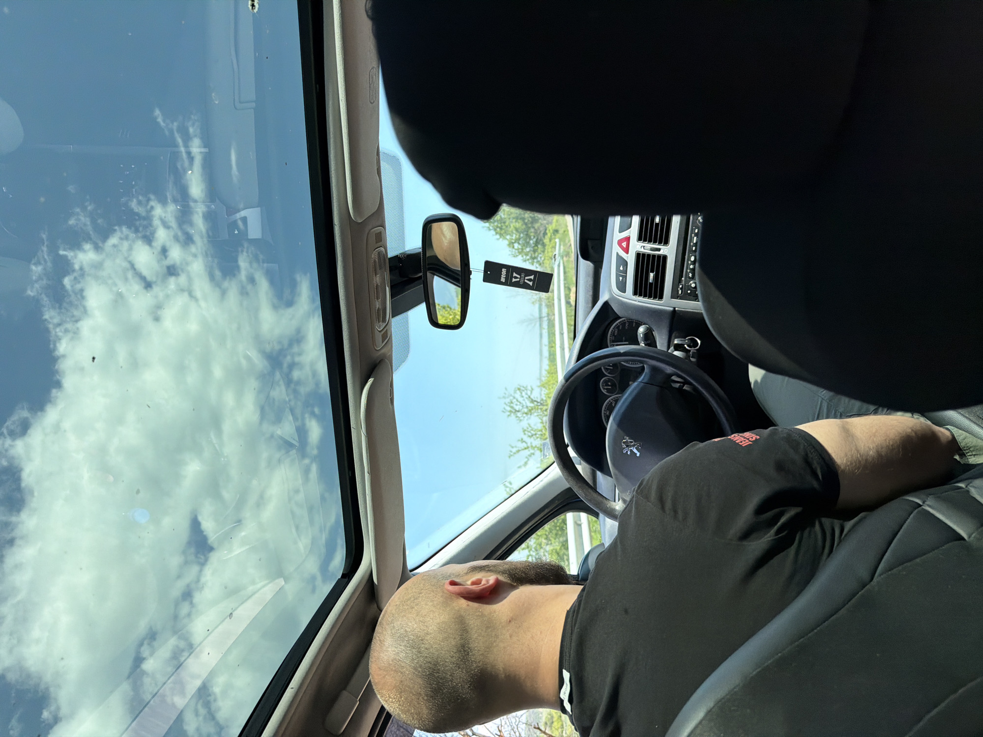 The image depicts the interior of a Peugeot car, likely in Belovitsa, Bulgaria. The view is from the passenger seat, angled towards the driver. The driver, a bald man with a black T-shirt, is either resting or asleep, as he is reclined towards the center console. The steering wheel prominently features the Peugeot logo. The car's dashboard and console are visible, along with an air freshener hanging from the rearview mirror. The sunroof is open, reflecting a cloudy sky. Outside, there are trees and greenery. The scene suggests the car is parked on a road.