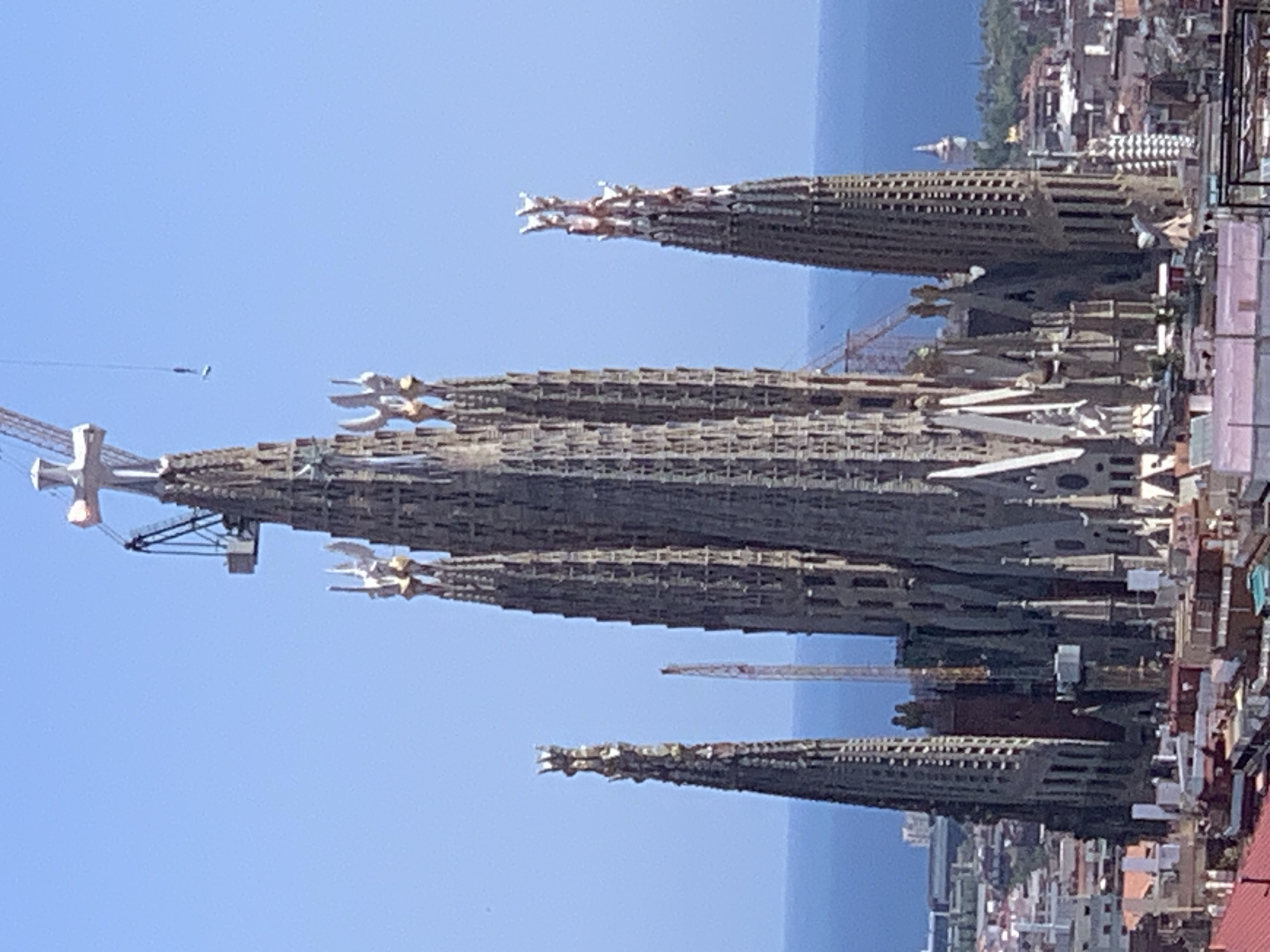 The image shows a close-up, aerial view of the Sagrada Familia basilica in Barcelona, Spain, under construction. The iconic spires of the basilica dominate the frame, with a prominent cross atop one of the tallest towers. Cranes are visible, one extending from behind the basilica and another with a hanging platform over the central section, indicating ongoing construction work. Below the basilica, a dense urban landscape of rooftops and buildings stretches towards the horizon, where the blue of the Mediterranean Sea is visible. The sky is clear and blue, suggesting a sunny day.