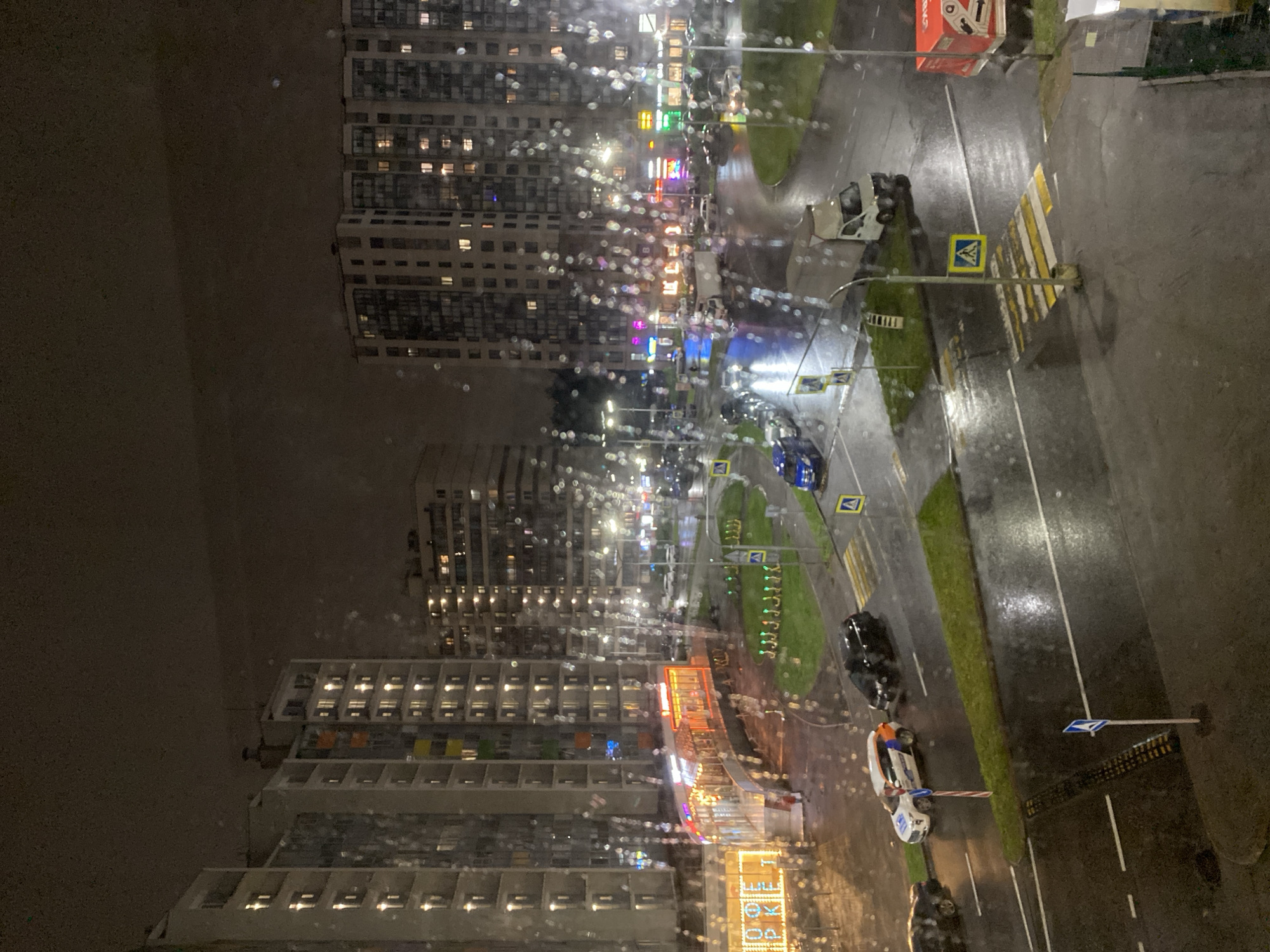 A high-angle, nighttime view shows a city scene in Unknown, Unknown. Two tall buildings with lit windows stand prominently on the left. A street runs vertically on the right side of the image, marked with lanes, pedestrian crossings, and traffic signs. Numerous cars are parked along or moving on the street. There is also a police car. Streetlights cast reflections on the wet asphalt, and rain droplets blur the view on the window from which the photo was taken.