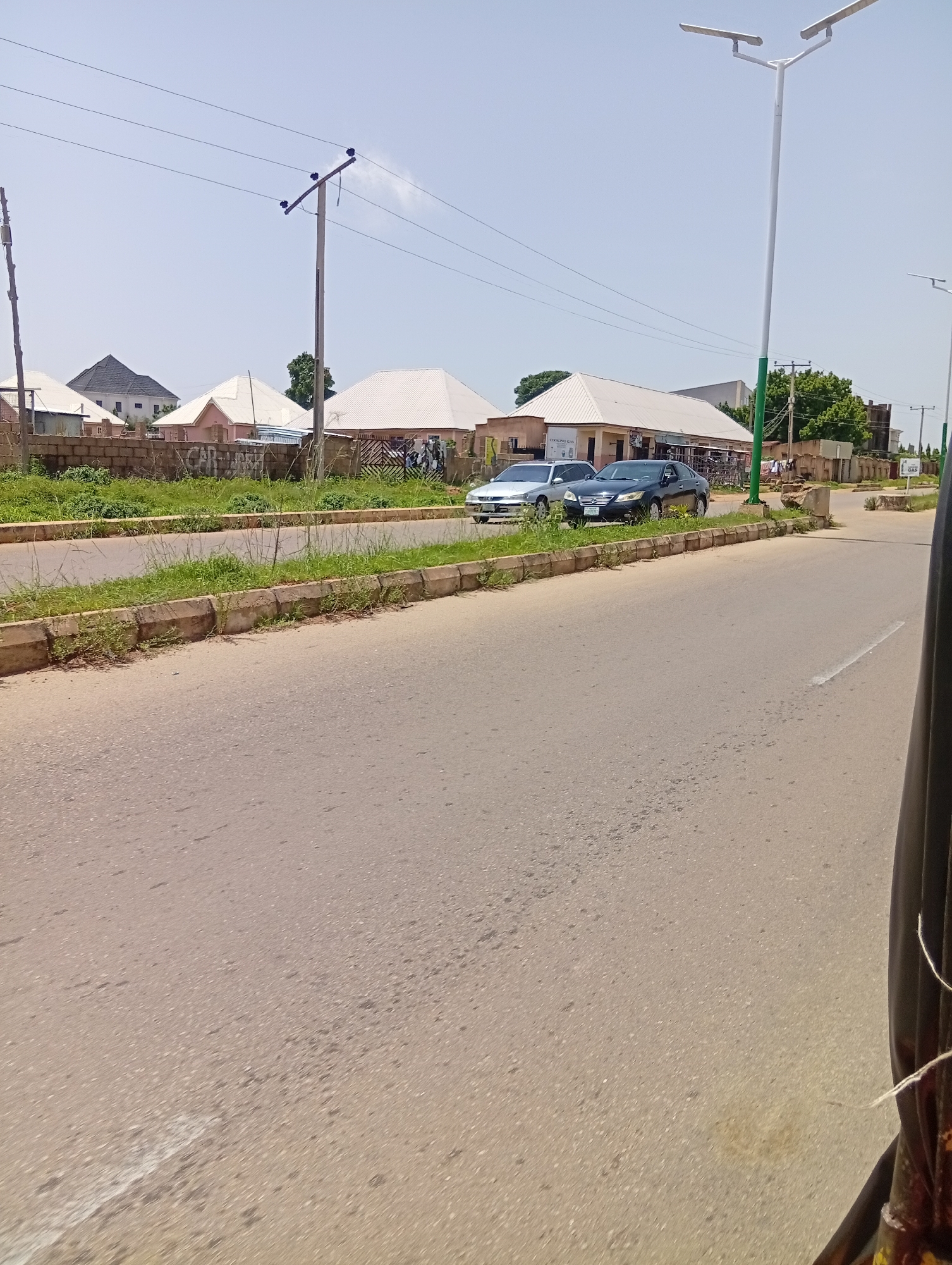 The image captures a daytime scene on a street in Jos, Nigeria. The road in the foreground is paved and appears to be a public thoroughfare. On the left side of the road, there's a strip of grass and vegetation bordered by a concrete curb, separating it from a wall and several buildings. These buildings have white, sloped roofs and appear to be shops or residences. In the background, there are utility poles with electrical wires stretching across the sky, indicating infrastructure for electricity.
Two cars are visible on the road. A silver sedan is parked on the left side of the road, and a dark sedan is parked slightly behind it. Beyond the cars, more buildings and some trees can be seen. A large, modern street lamp with two lights stands prominently on the right side of the road. The sky is clear and bright, suggesting a sunny day. The overall impression is of a typical urban or suburban street scene in Nigeria, possibly during business hours given the shops. There are no people prominently featured, and the general mood appears neutral and observational. No specific events or activities are clearly depicted, beyond the presence of parked cars on what seems to be a street. A small sign that says "GAS" is visible on the right side of the frame.