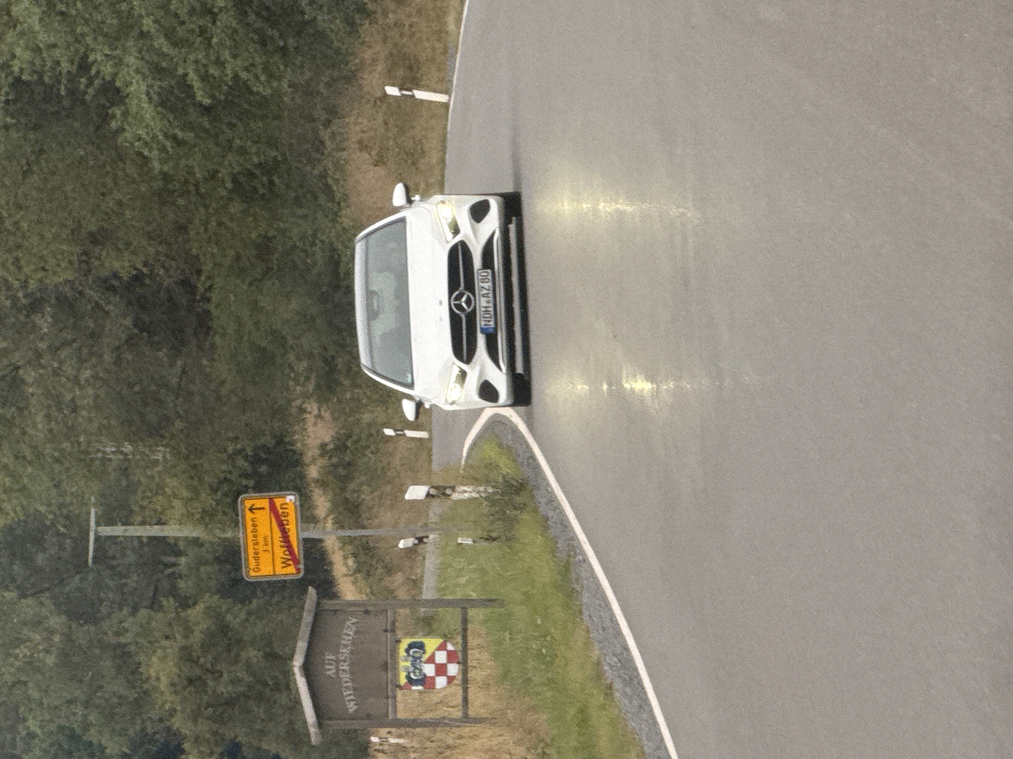 A white Mercedes-Benz A-Class car with the license plate "NDH-AZ 80" is driving on a wet, paved road. The car is in the foreground, facing the camera, and its headlights are on. To the left of the road, there is a wooden sign with the text "AUF WIEDERSEHEN" and a shield with a crest. Further to the left, a yellow road sign indicates directions to "Gudersleben" (1 km) and "Wolfleben" with a red line through "Wolfleben". The environment is rural, with trees and vegetation in the background. The scene appears to be in Germany, possibly near Ellrich, given the style of the road signs.