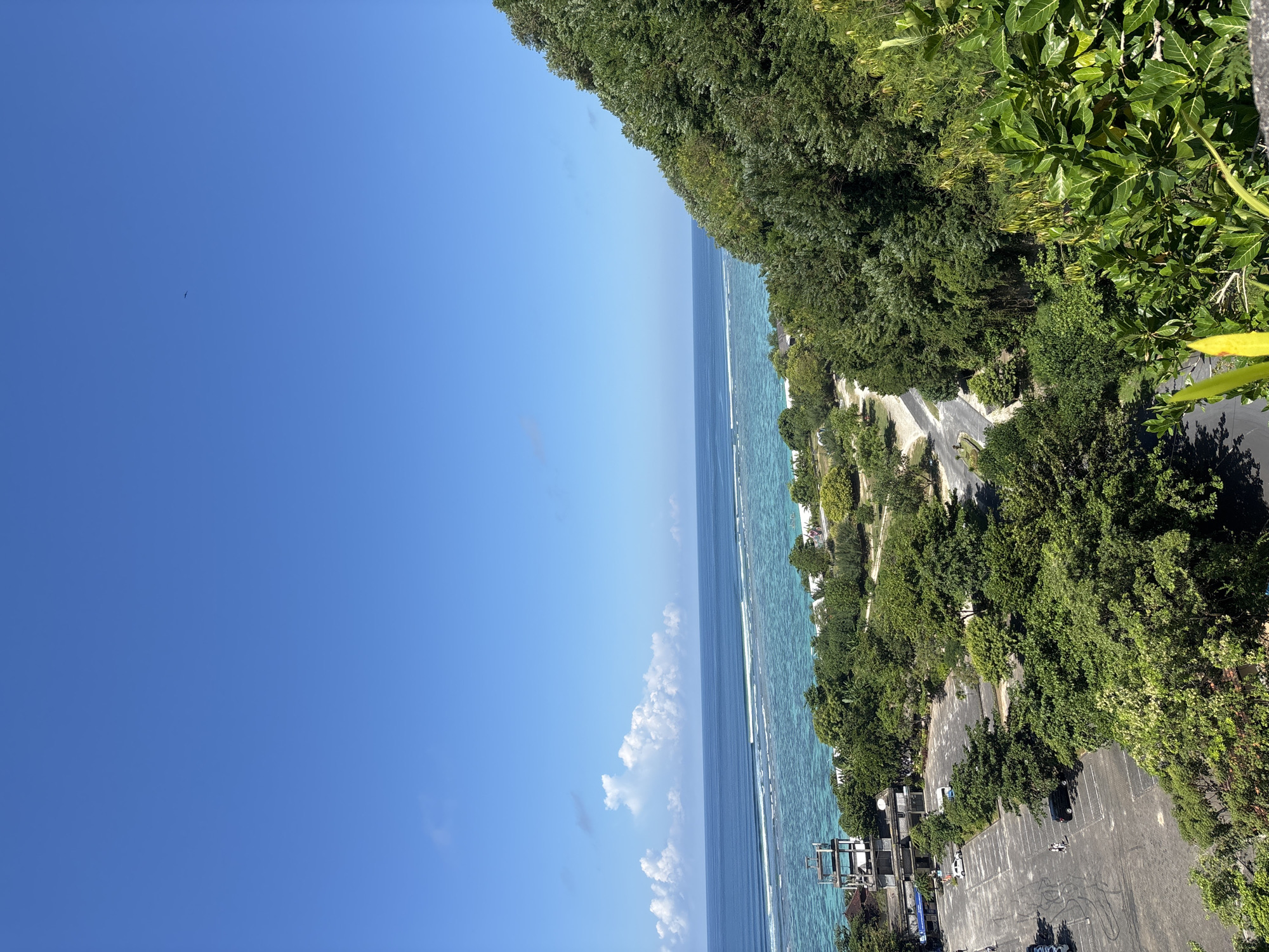 The image shows a coastline in Kutuh, Indonesia, from an elevated perspective. The scene features a deep blue sky with a few scattered clouds. A light blue ocean stretches alongside a coastal road with buildings and parking areas. Lush green trees cover the land bordering the road. A small bird is seen in the sky.
