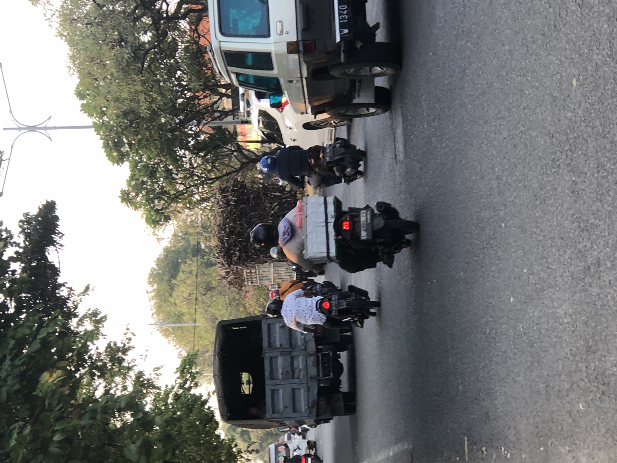 The image captures a street scene in Sumorame, Indonesia, with several vehicles and individuals navigating the road. A large, gray truck, possibly a military surplus vehicle, is prominent in the left-center of the frame, its cargo bed empty. Several motorcycles are present, with riders wearing helmets. One motorcycle in the foreground has a white storage box mounted on the back. To the right, a white SUV with black trim is visible, and a white van is seen in the distance on the left. Trees line the street, and the sky is overcast, suggesting either early morning or late afternoon light. The road surface appears to be asphalt.