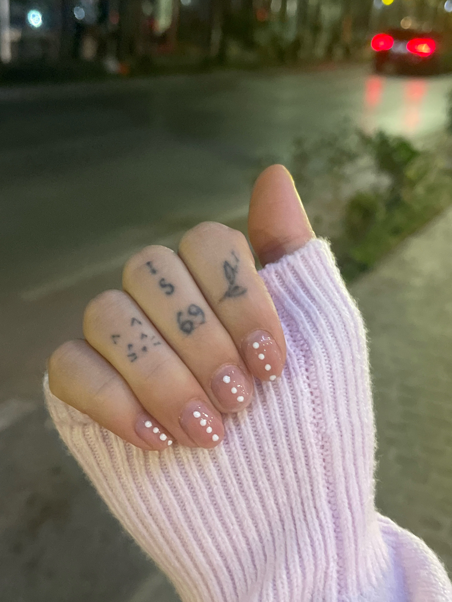 A close-up, nighttime, outdoor shot shows a person's left hand, which is wearing a light pink knitted sleeve. The hand is covered in small, dark tattoos and has light-colored nail polish with white polka dots. The background is blurred, showing out-of-focus streetlights and a vehicle with red taillights, suggesting a city environment.