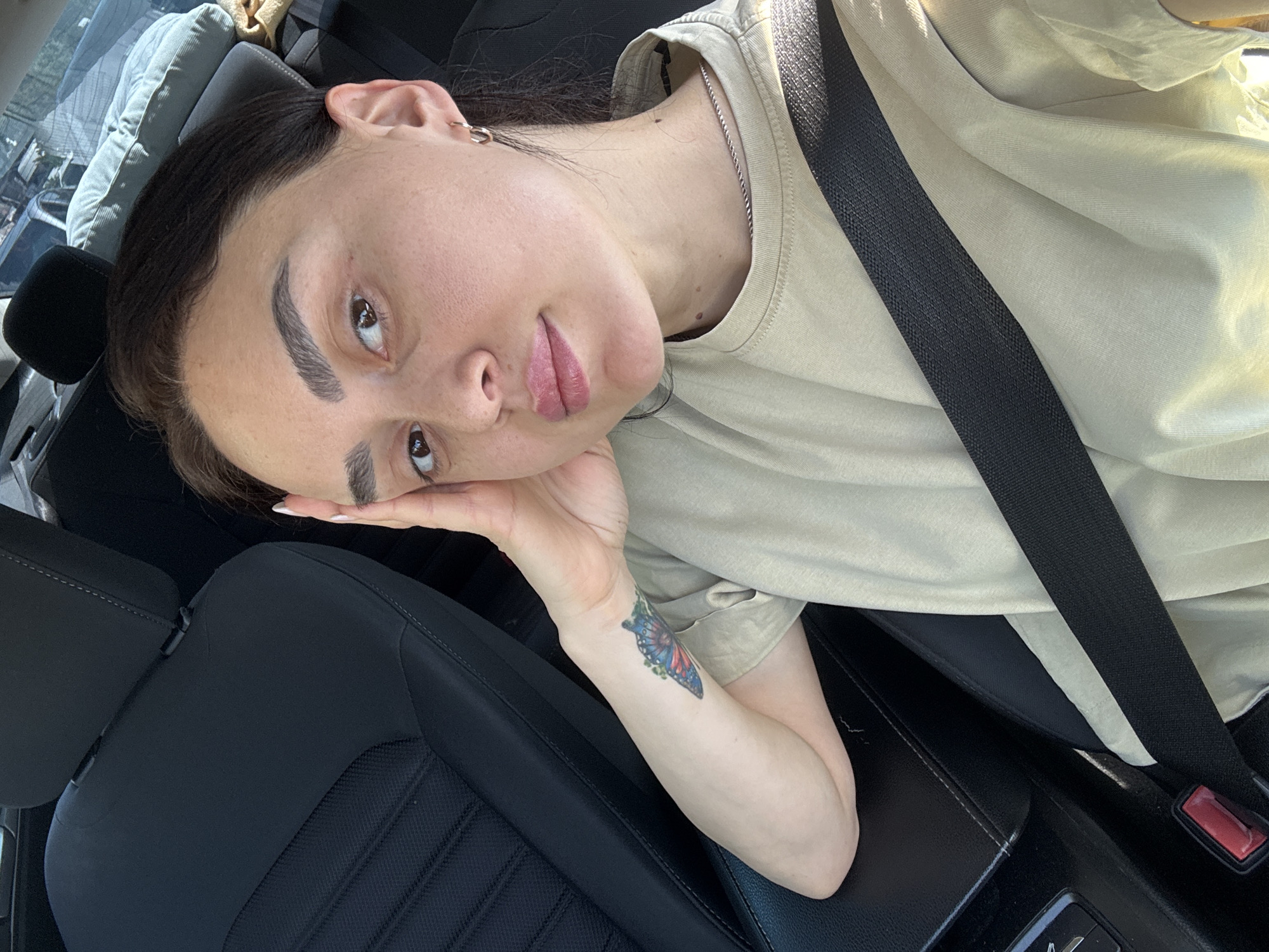A woman is seated in a car, wearing a seatbelt and a light tan t-shirt. She is leaning her head on her hand, which has a butterfly tattoo on the wrist. The car has black interior. The location is unknown; Tbilisi, Georgia, cannot be confirmed from the image.