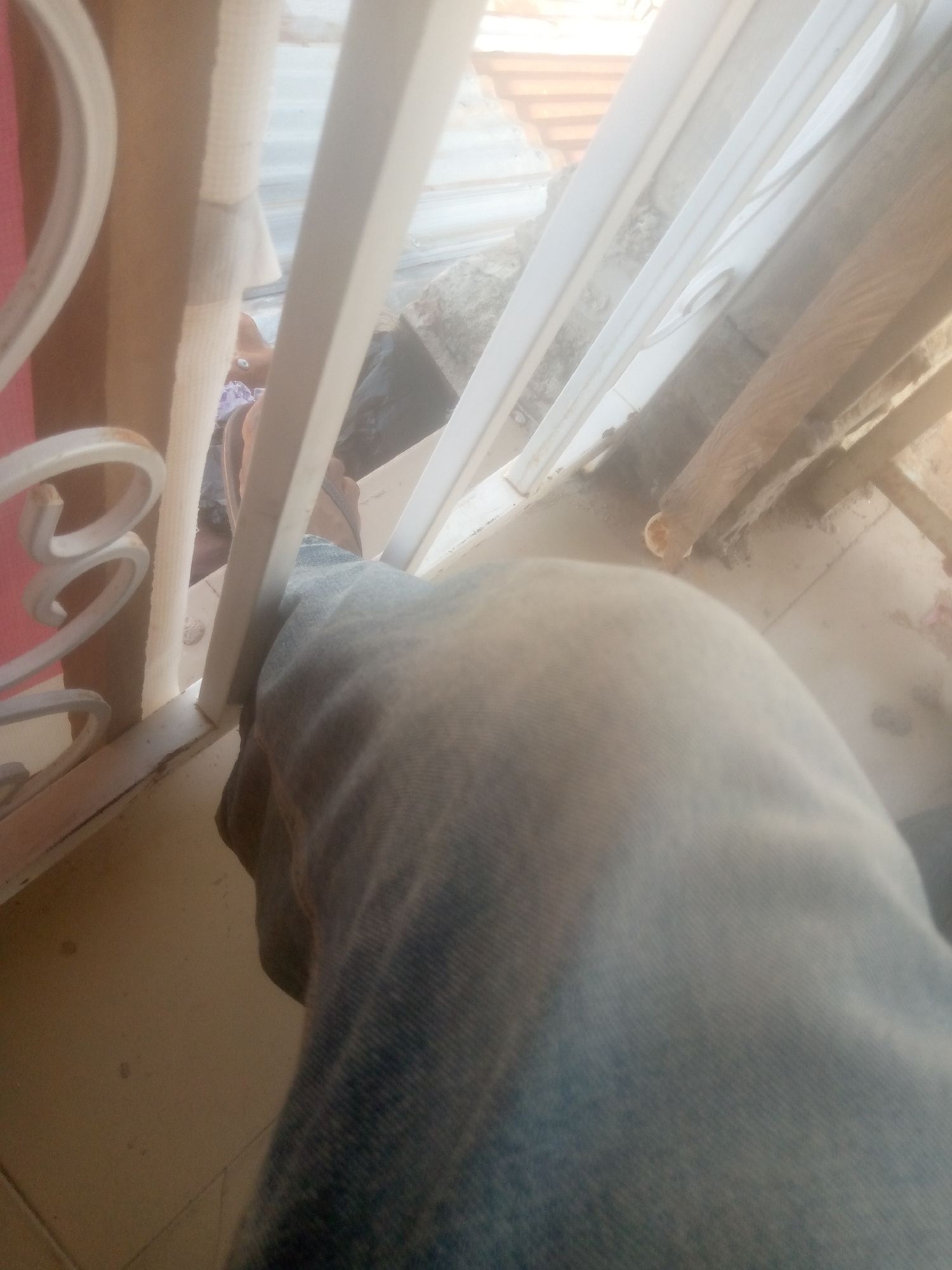 An outdoor scene, likely near a window or balcony, with a person's lower leg and foot visible. The person is wearing blue denim jeans and a sandal. The environment appears somewhat cluttered with visible corrugated roofing material and wooden beams in the background, suggesting an unfinished or outdoor construction area. The foreground is dominated by the person's leg and a beige tiled floor. The lighting is bright and somewhat overexposed, indicating daytime. This setting might be a residential area in Jalingo, Nigeria, potentially a balcony or an area under construction.