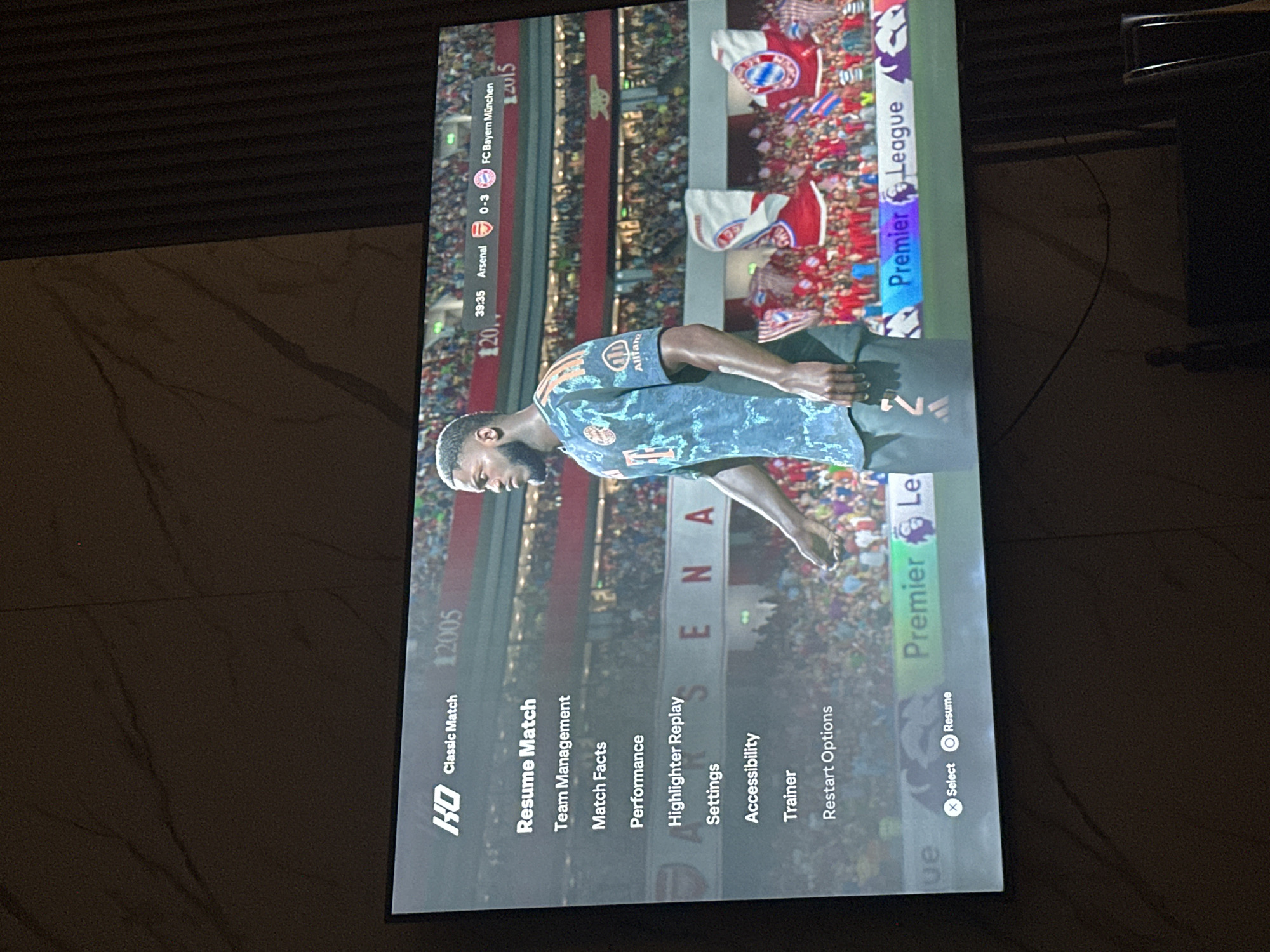 The image displays a television screen showing a football match simulation, likely from a video game, featuring a menu overlay. The game appears to be a classic match in the "HD" category, with the score indicating Arsenal 0-3 FC Bayern München at 39:35. The menu options visible include "Resume Match," "Team Management," "Match Facts," "Performance," "Highlighter Replay," "Settings," "Accessibility," "Trainer," and "Restart Options." At the bottom of the screen, the "Premier League" logo is visible, along with controller prompts for "Select" (X button) and "Resume" (O button). The scene is captured in a dimly lit room, with the television screen being the primary light source. The background of the room is dark and indistinct, suggesting it is nighttime or the lights are off. The context of Gwarinpa, Nigeria, is not directly depicted or implied by any visual elements within the image itself. The image is a close-up of a television, and no external environment is visible.