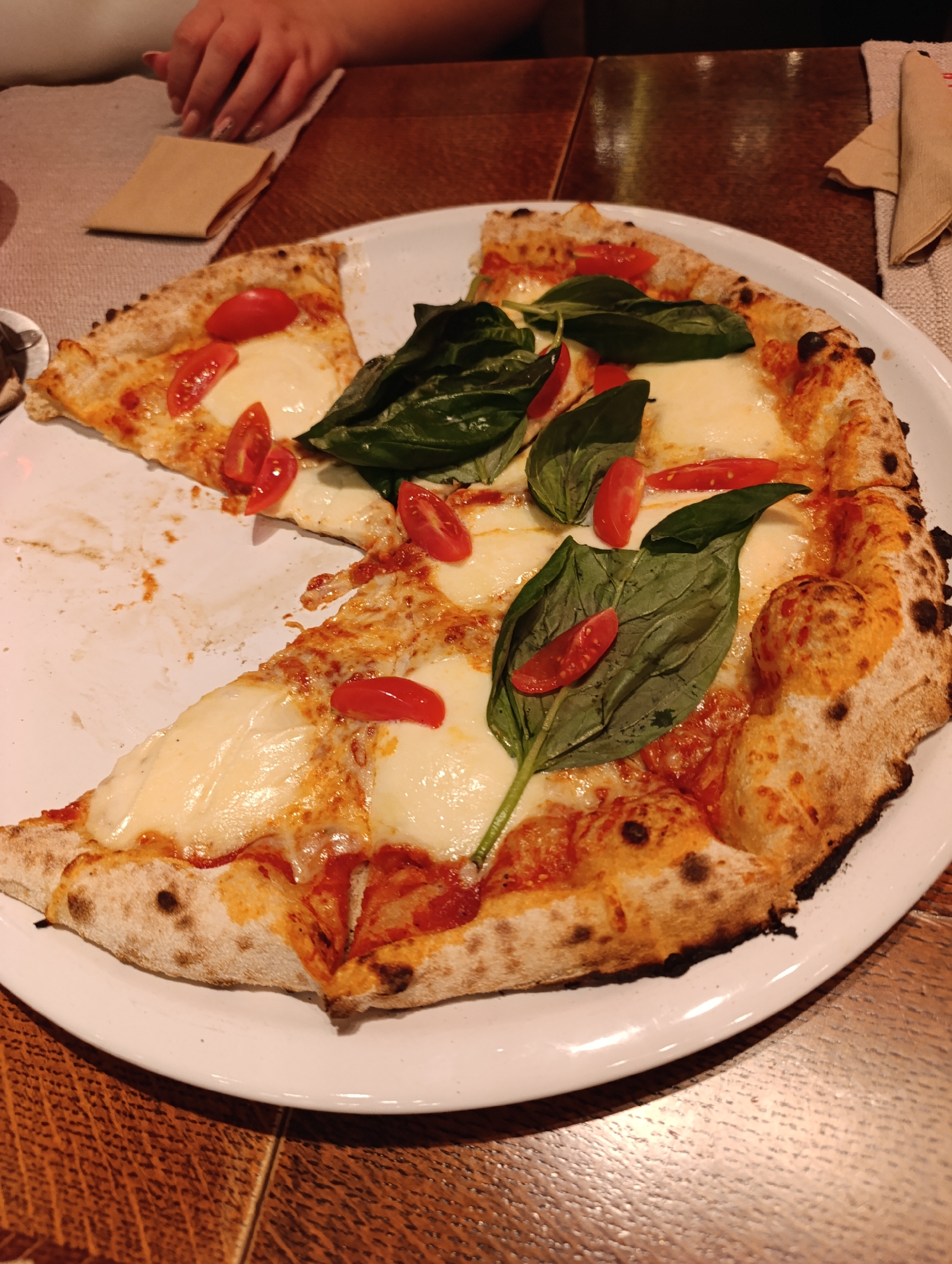 A partially eaten pizza sits on a white plate on a wooden table. The pizza is topped with mozzarella cheese, sliced tomatoes, and fresh basil leaves. A slice is missing from the pizza, and the plate has some crumbs and sauce residue. A folded napkin and a woman's hand are visible in the background. The environment suggests a restaurant setting. There is no information in the image to indicate the location is Haskovo, Bulgaria.