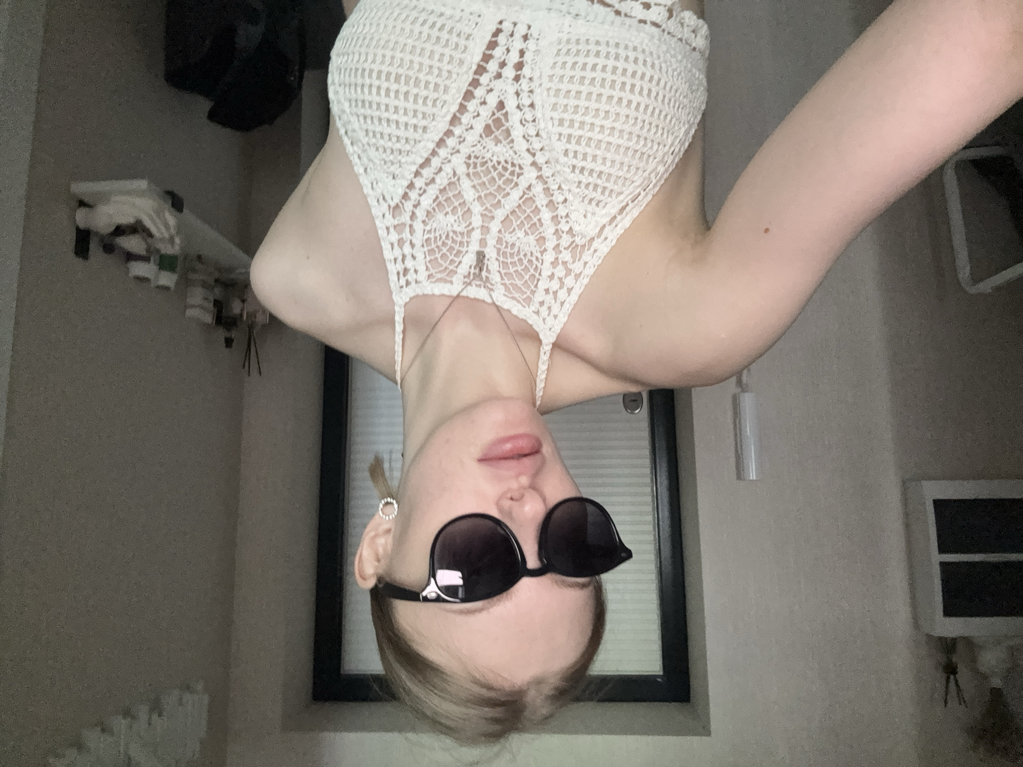 A person wearing sunglasses and a white crocheted top is photographed indoors. The person's body is oriented with the head downwards in the frame. The background features a window with closed blinds, a wall, and various items on a shelf. The location is unknown. The city of Perm, Russia is not visibly indicated in the image.