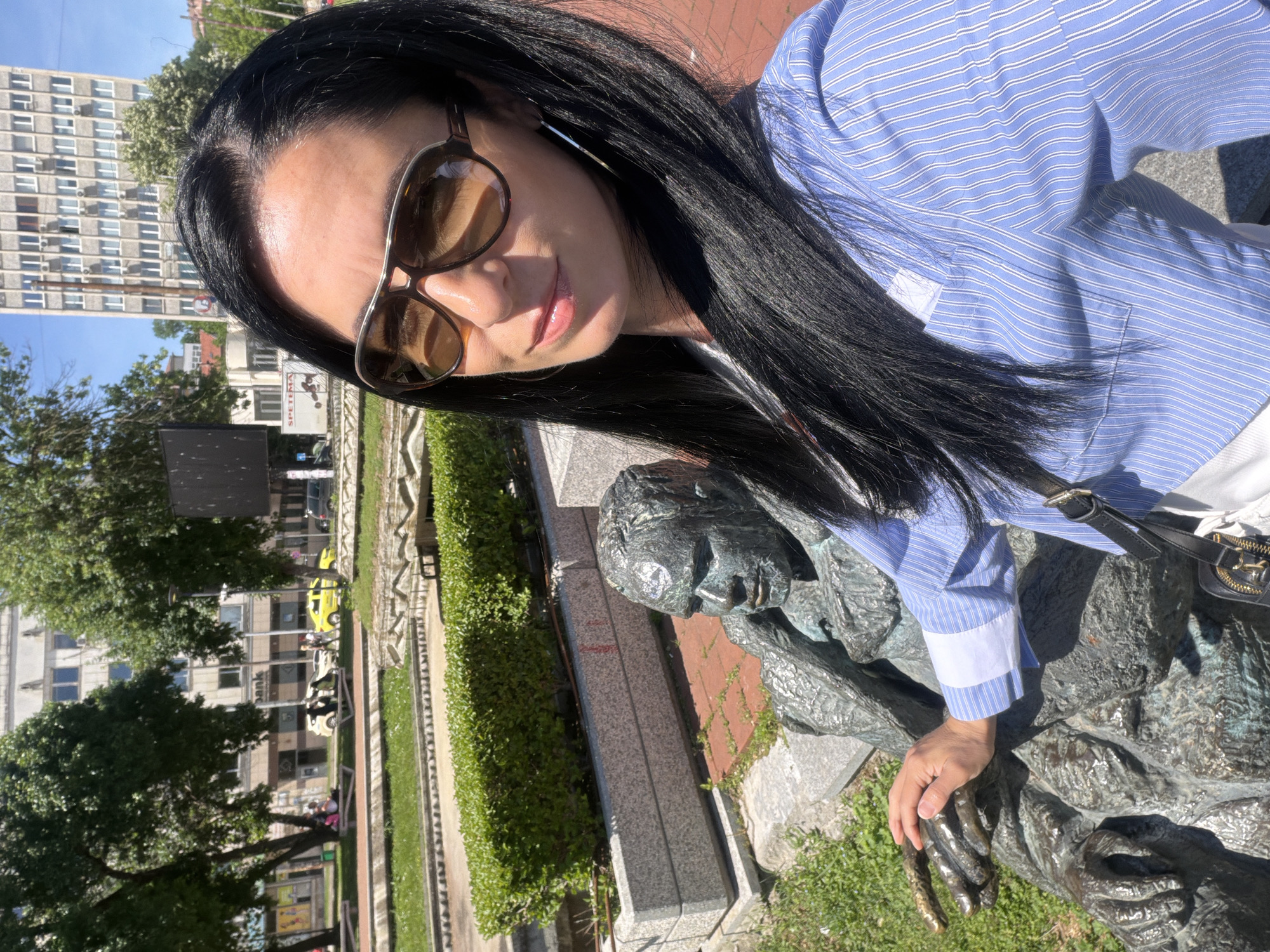 The image shows a woman posing for a selfie. She is wearing sunglasses and a blue striped shirt. Her black hair is long and straight. She is standing next to a bronze statue of a seated man in a thoughtful pose, her hand resting on the statue's hand.
The scene appears to be outdoors in a public space, possibly a park. A building and trees are visible in the background, along with parked cars and a city street. This is the statue of Peko Takov located in Pleven, Bulgaria.