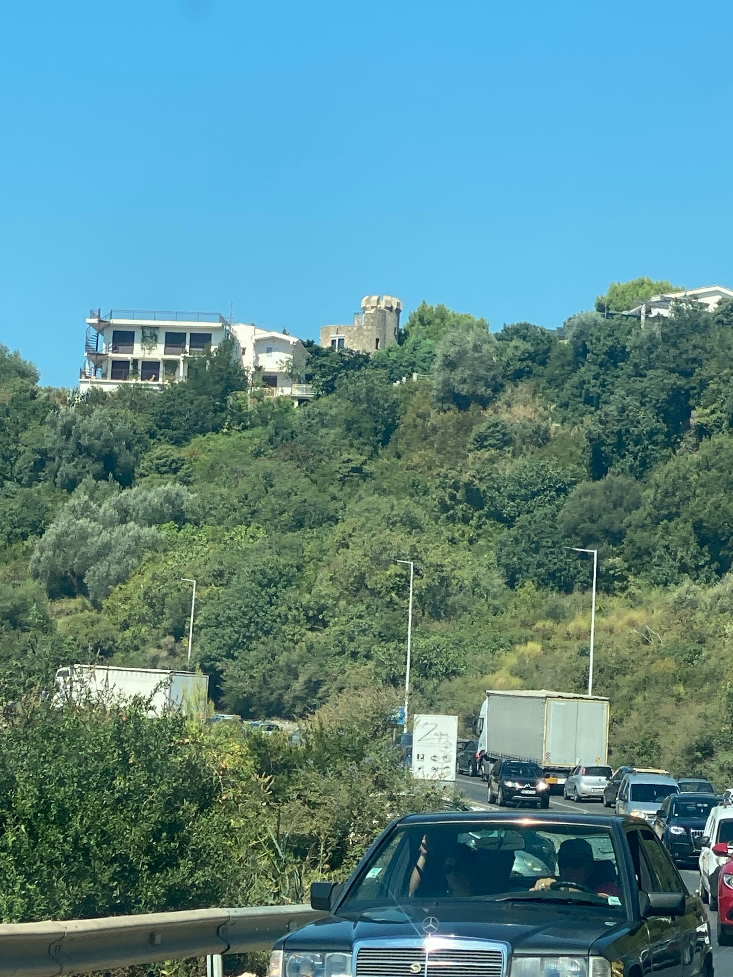 The image depicts a traffic jam on a sunny day, likely on a road leading into or out of Sutomore, Montenegro. In the foreground, a black Mercedes-Benz sedan is stopped, with two individuals visible in the driver and passenger seats. The road ahead is congested with numerous cars, including a silver minivan, a black SUV, and several other vehicles. Further back, a large white box truck is visible, also part of the traffic. The background features a lush, green hillside covered in dense vegetation. Several buildings are scattered across the hillside, including a multi-story white building with balconies and a spiral staircase, and a stone tower-like structure. The sky is clear and blue.