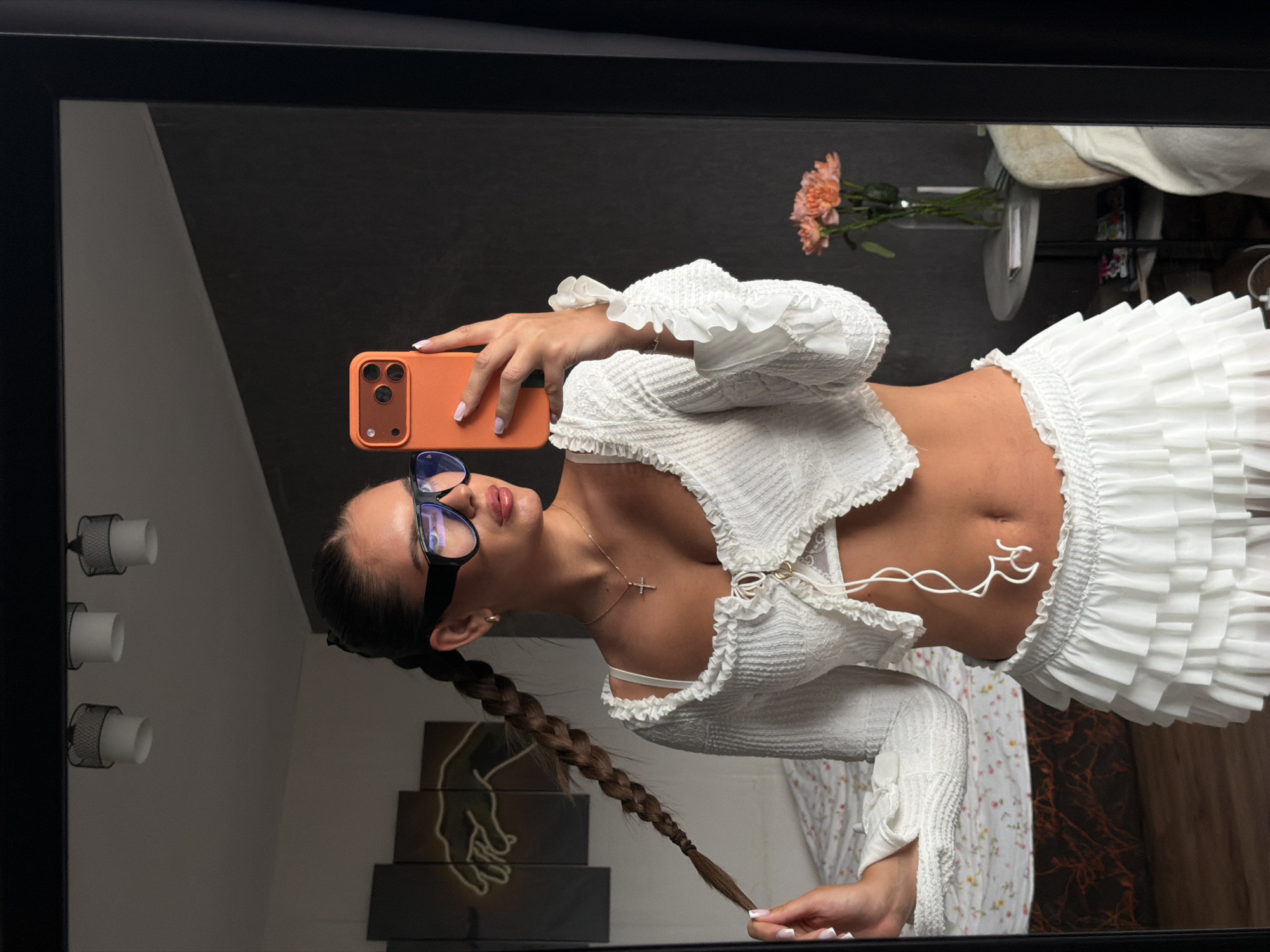 A woman with fair skin and dark hair braided and pulled back is posing for a mirror selfie. She is wearing a white ruffled top and a matching white mini skirt with multiple layers of ruffles. The woman is wearing glasses with thick black frames and a gold necklace with a cross pendant. She is holding an orange smartphone with a triple-lens camera.
The reflection in the mirror reveals a room with a black wall and a white wall on the left. There is a light fixture with three cylindrical lights on the left side of the mirror, and a vase with orange flowers on the top right. A stack of boxes with a hand-drawn silhouette on top is visible near the bottom left of the mirror. A floral-patterned robe and a light-colored drapery are seen in the lower part of the reflection.
There is no information given that this image was taken in Krasnoyarsk, Russia.