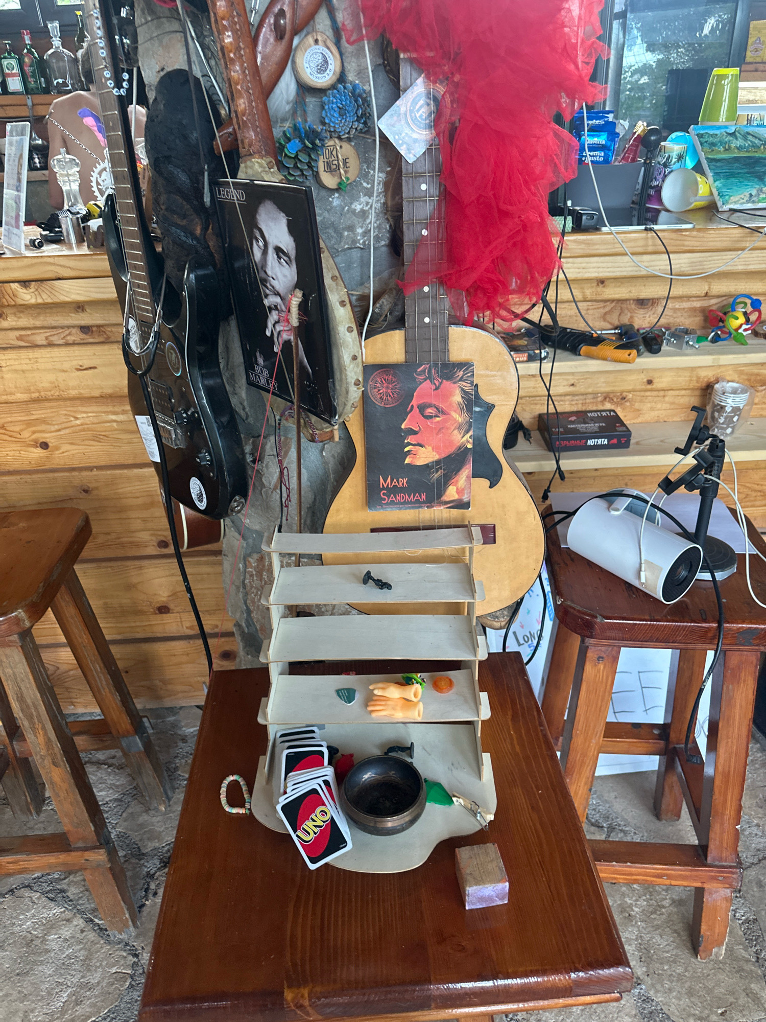 The image depicts an indoor, rustic-themed setting that appears to be a personal collection or display area, possibly in Cetinje, Montenegro, given the presence of traditional wood construction and decorative elements. The background consists of rough-hewn wooden walls. Prominently featured are several guitars, some hanging vertically and others propped up. One guitar has a poster of Mark Sandman attached to its body.
In the foreground, a tiered shelf unit holds various small objects, including what appear to be game pieces, a small orange figure, and a toy hand. Below the shelf, a deck of UNO cards and a small metal bowl are visible on a wooden table. To the right of the table, a wooden stool supports a white cylindrical object with a black stand, likely a speaker or projector, connected by wires.
The overall impression is one of a lived-in space filled with personal interests, with a strong emphasis on musical instruments and assorted decorative items. The lighting suggests an indoor environment, possibly with natural light filtering in from an unseen source. There are no people visible in the scene.