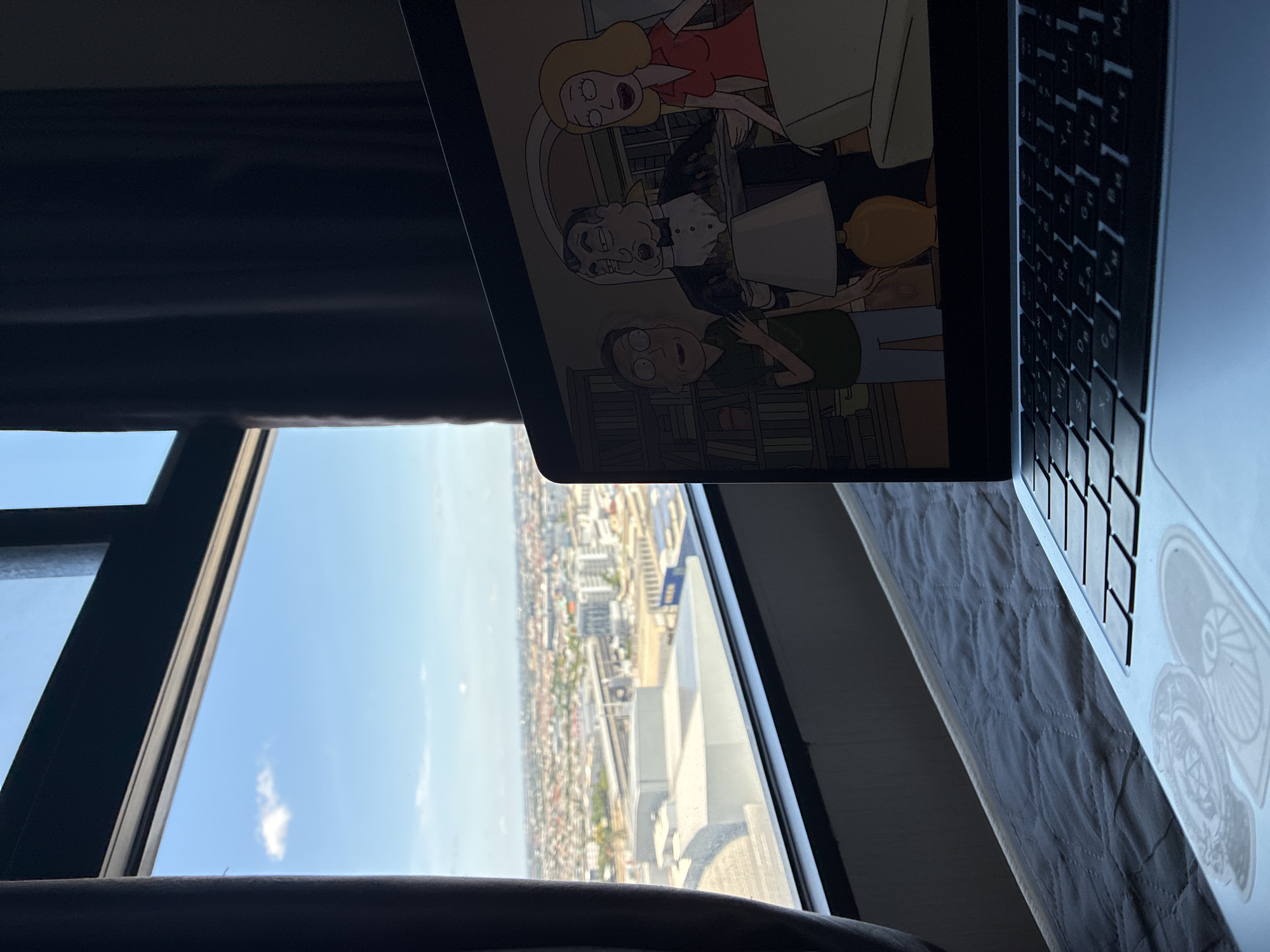 This media file depicts an indoor scene overlooking a city skyline. A laptop is open and positioned on a surface, likely a bed due to the quilted texture visible. The laptop screen displays an animated scene featuring characters from "Rick and Morty," specifically Summer, Rick, and Morty, engaged in conversation. A woman with blonde hair wearing a red dress stands, while Rick and Morty, in their signature green shirt and blue pants respectively, are seated.
The background of the laptop screen shows a bookshelf. To the right of the laptop, its keyboard is visible, with Thai characters on some of the keys indicating a Thai keyboard layout.
The laptop is placed near a large window, which offers a panoramic view of a densely populated city. The cityscape is characterized by numerous buildings, some appearing to be apartment complexes, and a train track cutting through the urban landscape. The sky is blue with a few scattered clouds, suggesting it is daytime. The angle of the sun, casting shadows and highlighting parts of the city, implies it is likely late morning or early afternoon.
The overall setting suggests a comfortable, perhaps residential, environment with a view of urban life. The presence of the laptop with a cartoon playing suggests a moment of leisure or relaxation. The time of day appears to be sunny and clear. The location is identified as Sao Thong Hin Town Municipality, Thailand, which aligns with the presence of Thai characters on the keyboard.