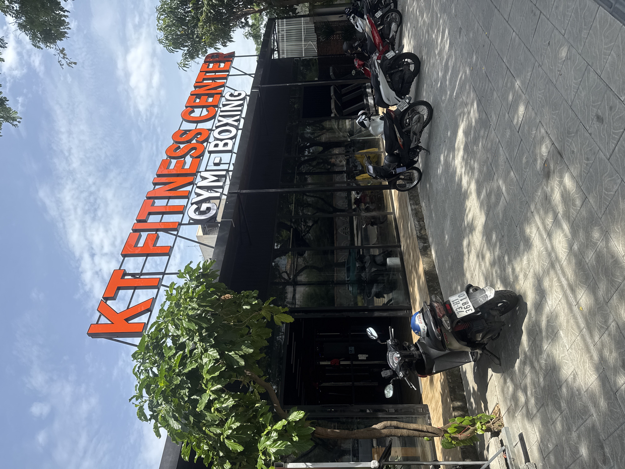 The image shows the KT FITNESS CENTER GYM & BOXING in Đà Nẵng, Vietnam. The building has a modern glass front. Several motorbikes are parked along the sidewalk in front of the building. A small tree grows near the entrance. The sky is visible, with light clouds.