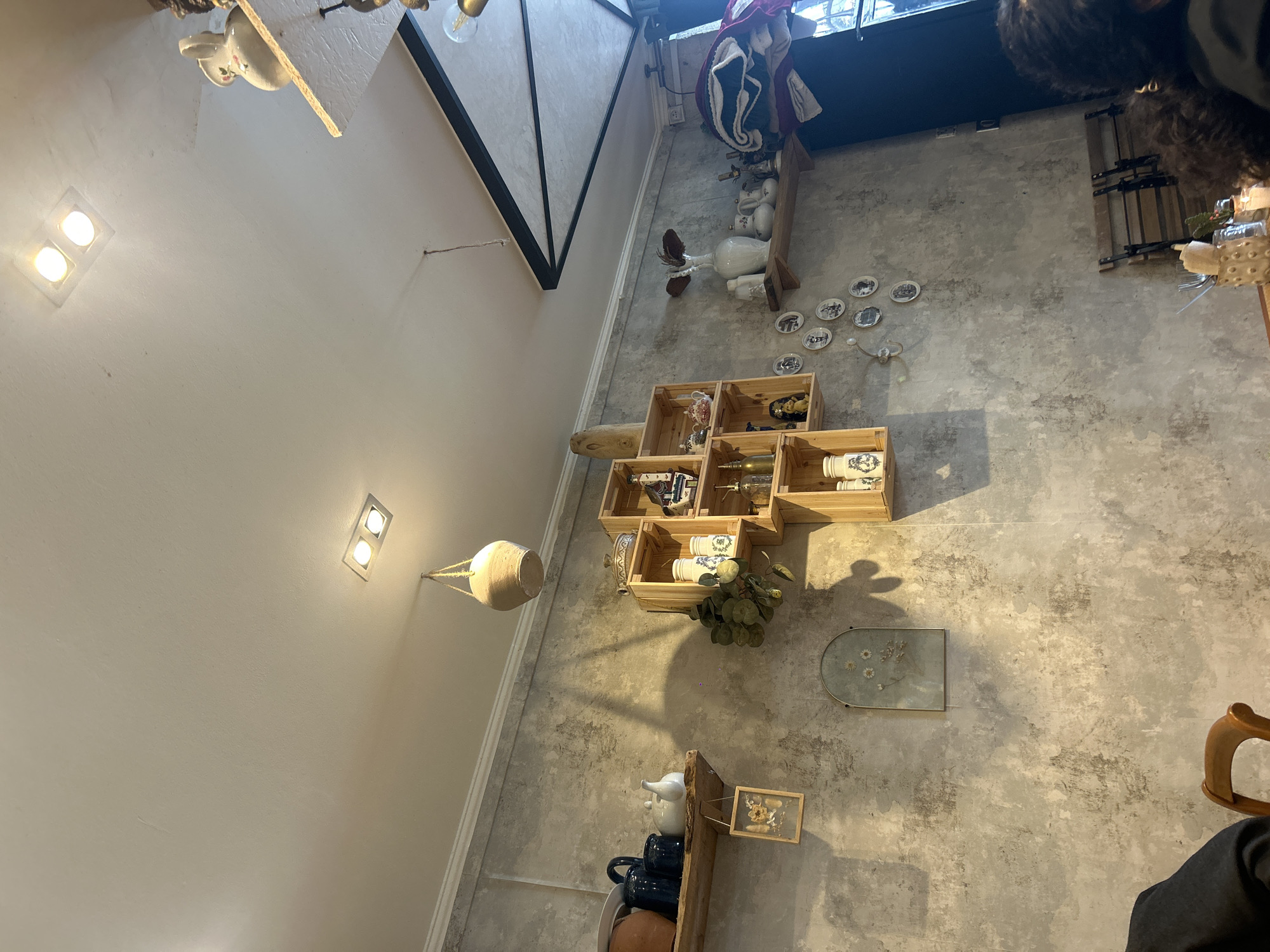 A high-angle shot shows a section of a wall with decorations inside a shop, possibly in Paris, France. The wall, seemingly concrete or stucco, features a collection of objects including wooden crates filled with white ceramic items, possibly vintage or antique pieces, and a framed botanical print. The arrangement is somewhat chaotic, with various objects placed at different heights and angles. A suspended woven basket hangs from the wall to the left. A portion of a person's head is visible in the upper-right corner of the shot.