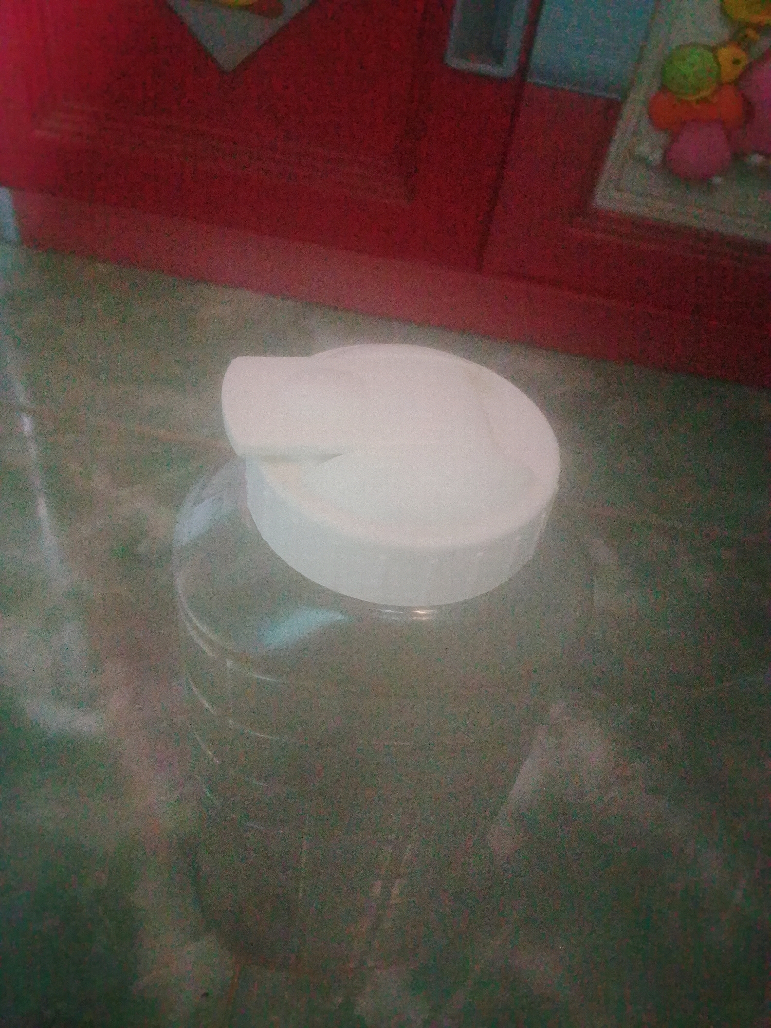 The image shows a close-up of a clear plastic bottle with a white lid, filled with a dark liquid. The bottle is placed on a textured, mottled surface that appears to be a tabletop or floor. In the background, a red surface with a textured pattern is visible. To the right of the red surface, a colorful object with round shapes is partially visible, possibly a toy or decoration. The lighting is dim, and the image has a grainy quality. There are no people or identifiable landmarks present in the image that would indicate a specific location in Kecamatan Haurgeulis, Indonesia.