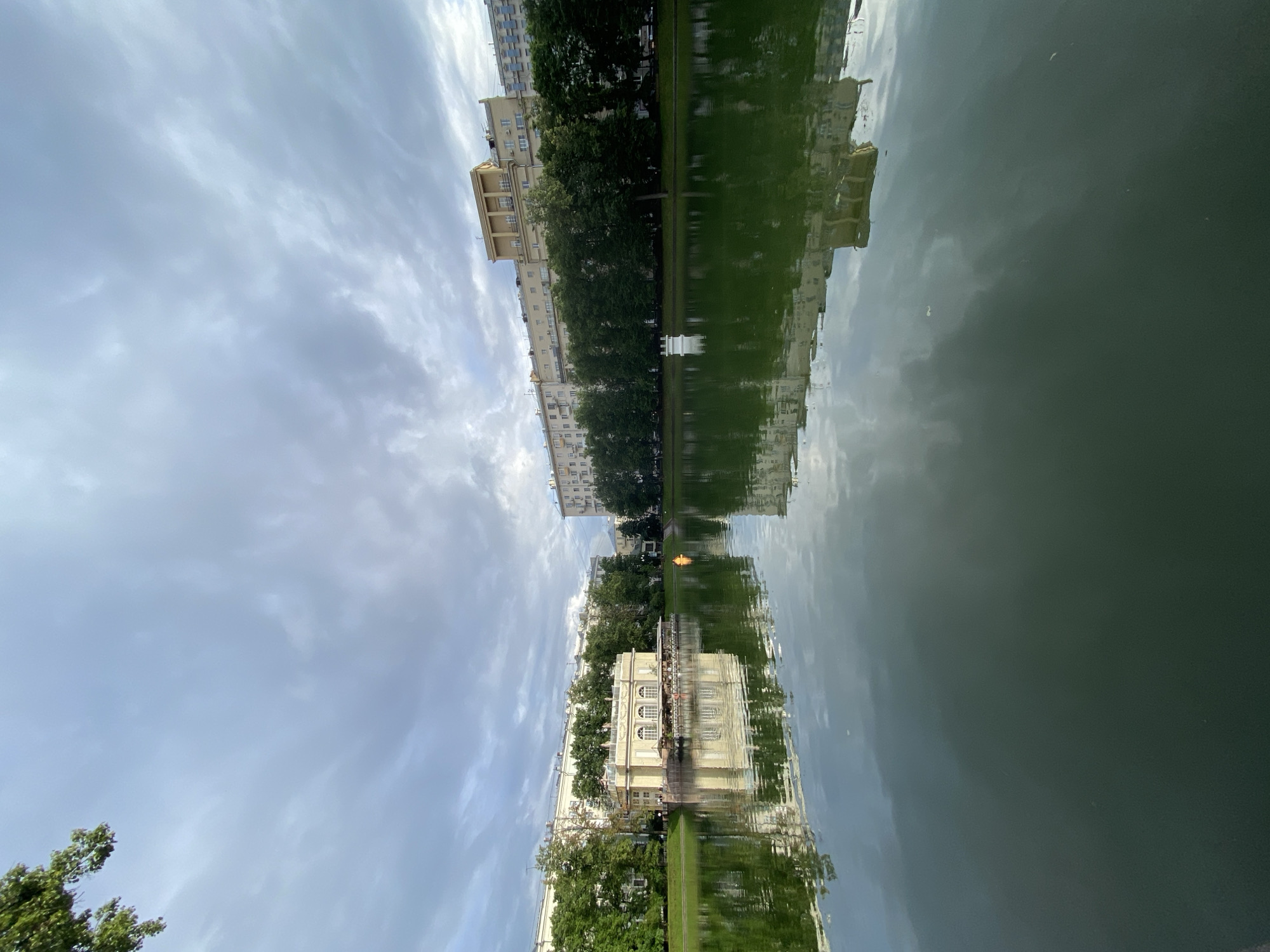 The image depicts a waterfront scene, likely a park or public space, with buildings and trees reflected in a body of water. The perspective is unusual, as the image is rotated, making the sky appear on the left and the water on the right, with the reflections forming the central and lower portions of the frame.
The foreground, which is actually the upper part of the original image, shows a cloudy sky. Below this, a line of tall, multi-story buildings with windows and balconies runs along what would be the edge of the water. Between the buildings and the water are lush green trees. The reflection in the water shows a mirror image of these buildings and trees. In the water, a separate, lighter-colored building with arched windows and an outdoor seating area is visible, also reflected. There appears to be a small, orange object in the water, possibly a buoy or a decorative element.
The overall mood is serene and perhaps a little disorienting due to the rotated perspective. The lighting suggests it is daytime, with diffused light due to the overcast sky. The presence of trees and a body of water indicates an outdoor, natural or semi-natural setting. Given the location context of Moscow, Russia, this could be a canal, river, or pond within a city park or historic area. No visible text is present in the image.