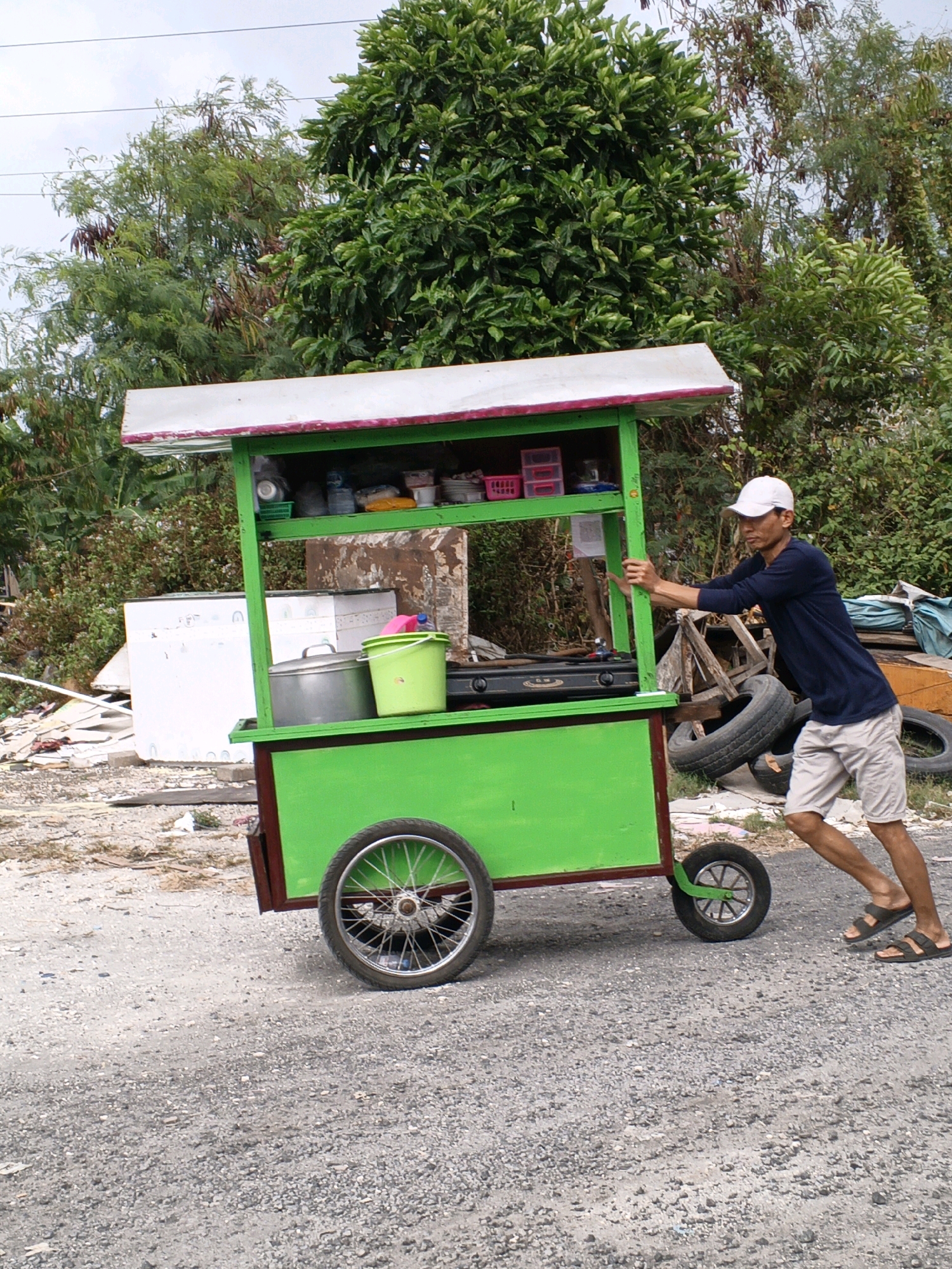 A man pushes a brightly painted green food cart on a gravel road. The cart has a white and pink striped canopy and is stocked with cooking equipment, pots, and buckets. The man, wearing a blue long-sleeved shirt, white cap, and beige shorts, is pushing the cart from the side. In the background, there is lush green vegetation and some discarded items, suggesting an outdoor setting on the outskirts of Denpasar, Indonesia.