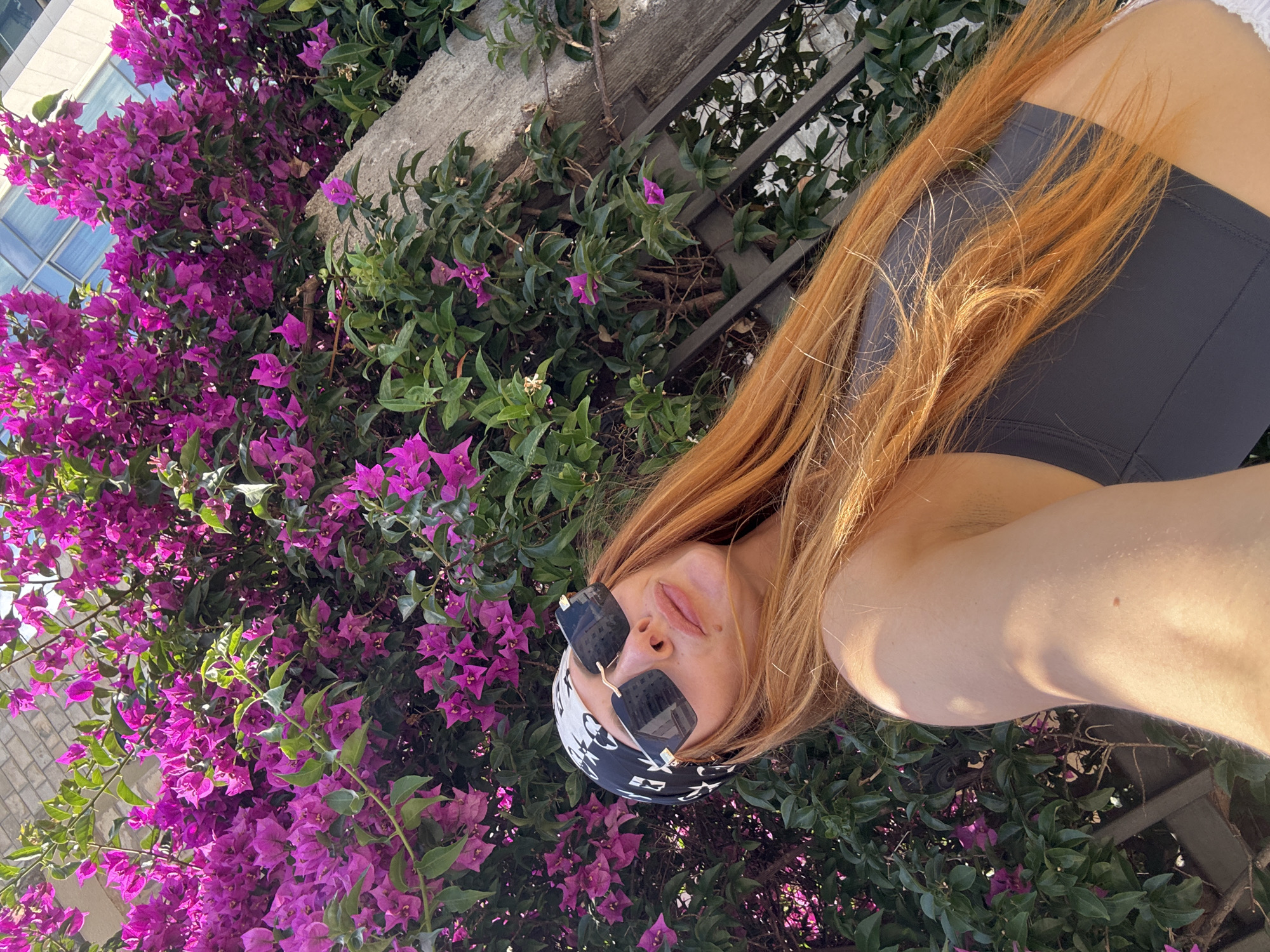 A young woman with long, reddish-blonde hair and sunglasses is taking a selfie outdoors, in front of a bush with vibrant purple flowers. She is wearing a black bandana with a white pattern and a dark grey sports bra. The background shows a brick wall and a modern building with reflective windows, suggesting an urban setting. The woman is positioned in the lower-left quadrant of the image, with her face turned towards the camera. The overall scene evokes a sense of warm weather and leisure.