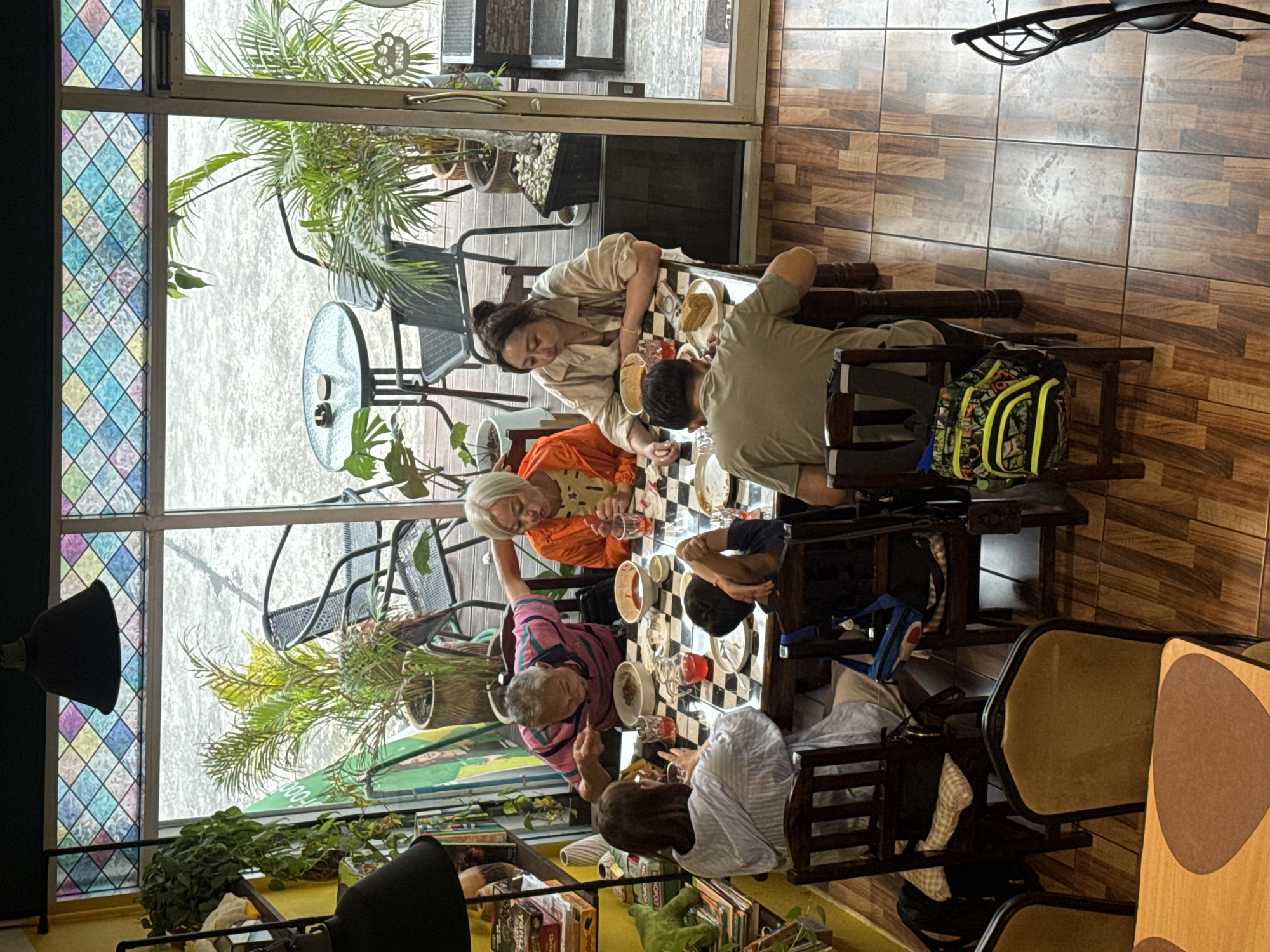 This image depicts a group of six individuals dining at a checkered-patterned table in what appears to be a cafe or restaurant. The scene is set indoors with large windows revealing an outdoor area with plants and outdoor seating. The individuals are engaged in eating and drinking. One man in a pink and blue striped shirt is actively eating from a bowl. A woman in an orange top is also eating. Another woman in a light-colored shirt is looking down at her food, and a person in a green shirt is seated with their back to the camera. Two children are also present at the table, one with their arm raised. The environment includes indoor plants, chairs, and decorative stained-glass panels above the windows. Based on the presence of outdoor seating, lush greenery, and the overall ambiance, this establishment is likely located in Chiang Mai City Municipality, Thailand, potentially a cafe that caters to both indoor and outdoor dining experiences.