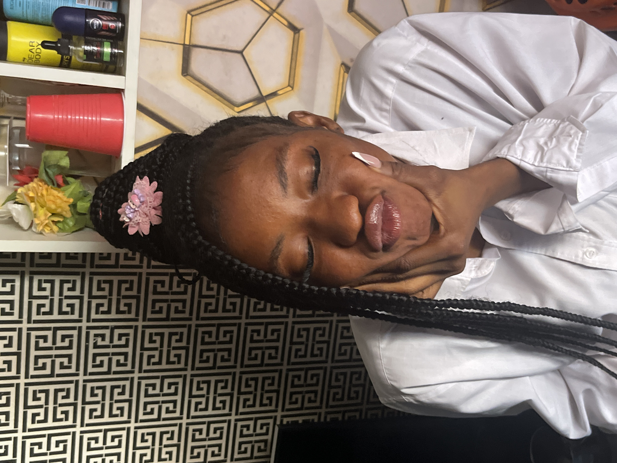 The image shows a close-up, sideways view of a young Black woman lying down, with her eyes closed and a peaceful expression. She is wearing a white button-down shirt, and her braided hair is adorned with a pink flower. Her lips are glossy, and a white nail polish is visible on one finger resting on her cheek. The background reveals a shelf with various objects, including red cups, and decorative geometric wallpaper. The lighting appears to be indoor, and there are no external weather cues. The overall mood is serene and relaxed. The visible text on the shelf includes "DEAR BODY" and what appears to be a brand name on a blue bottle, possibly a cosmetic or hygiene product. The provided location context is Jos, Nigeria.