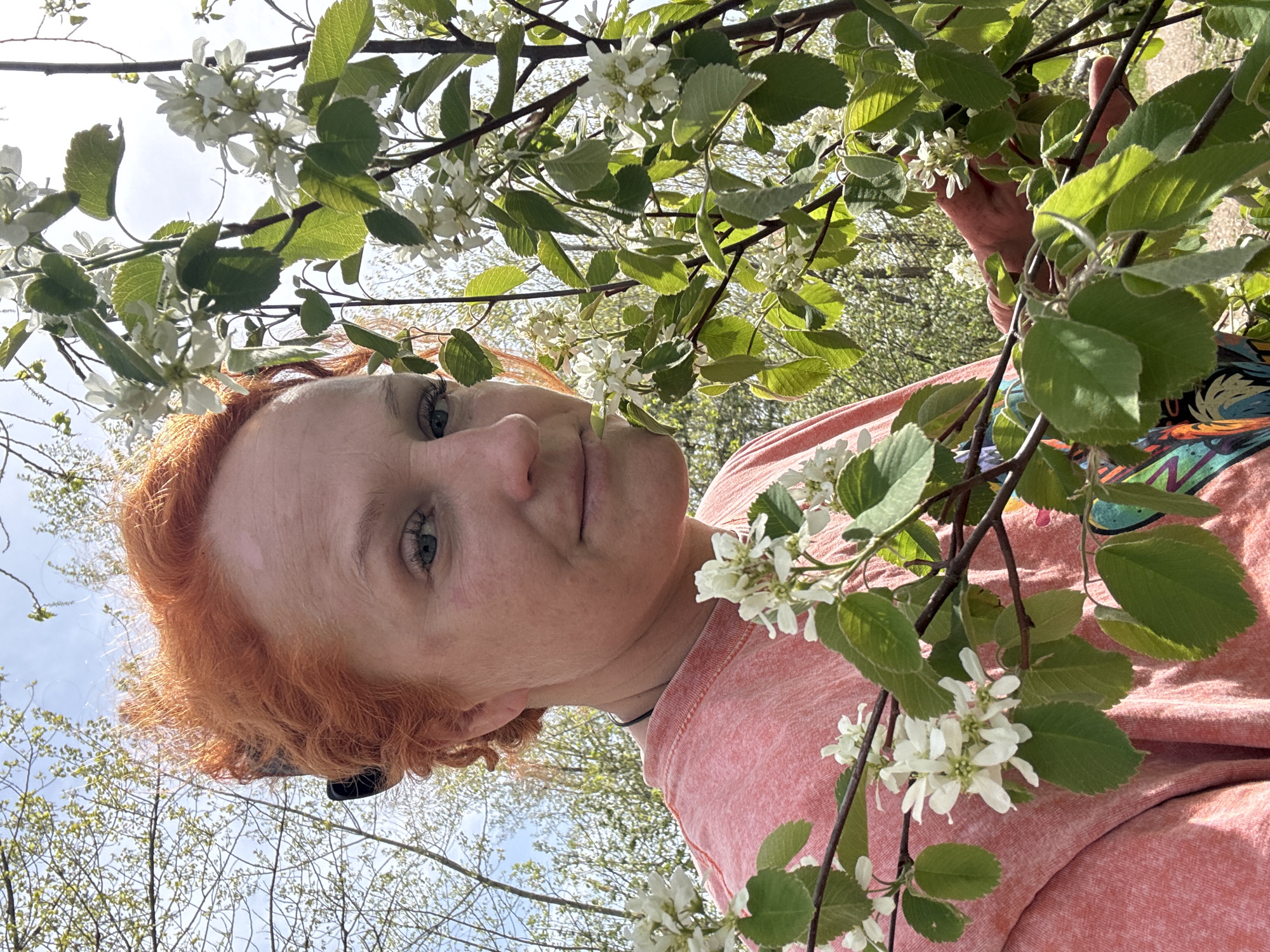 A woman with red hair stands outdoors, partially framed by branches with white blossoms and green leaves. She looks toward the camera with a neutral expression. The background suggests an outdoor setting with trees and a light blue sky visible through the branches. The woman is wearing a pink shirt. The location is unknown.