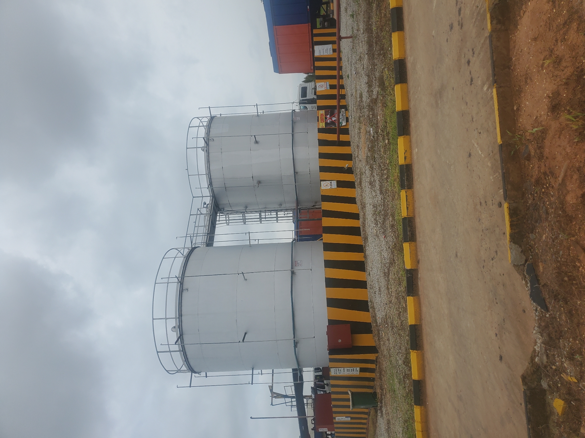The image shows an industrial scene in Gwagwada, Nigeria. Two large, cylindrical storage tanks, painted silver, are prominently featured. These tanks are connected by a walkway and a ladder system. They are surrounded by a black and yellow striped barrier, indicating a hazardous or restricted area. A green garbage bin is visible near the left side of the tanks. Several caution signs are posted on the barrier. In the background, a semi-trailer truck with an orange and blue container is parked. The sky is overcast, suggesting a cloudy day. The ground around the tanks is covered with gravel and some sparse vegetation. A paved road or pathway is in the foreground, with yellow and black striped curbing.
