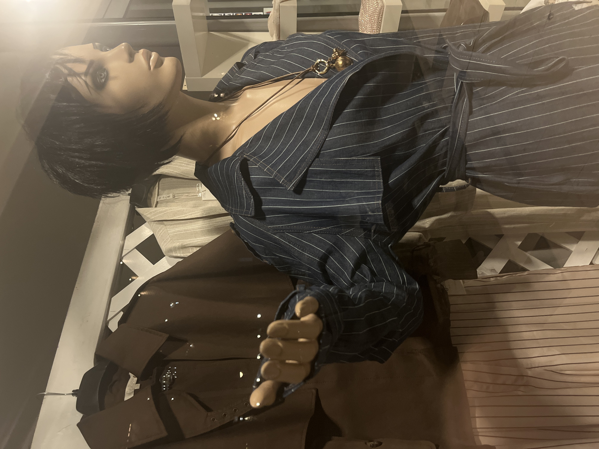 The image shows a mannequin in a storefront. The mannequin has short, dark hair and is wearing a striped navy blue shirt with a gold necklace. It's positioned among other clothes, including a brown garment with metallic details. The location is likely a retail establishment, but no specific identifiers point to Budva, Montenegro.