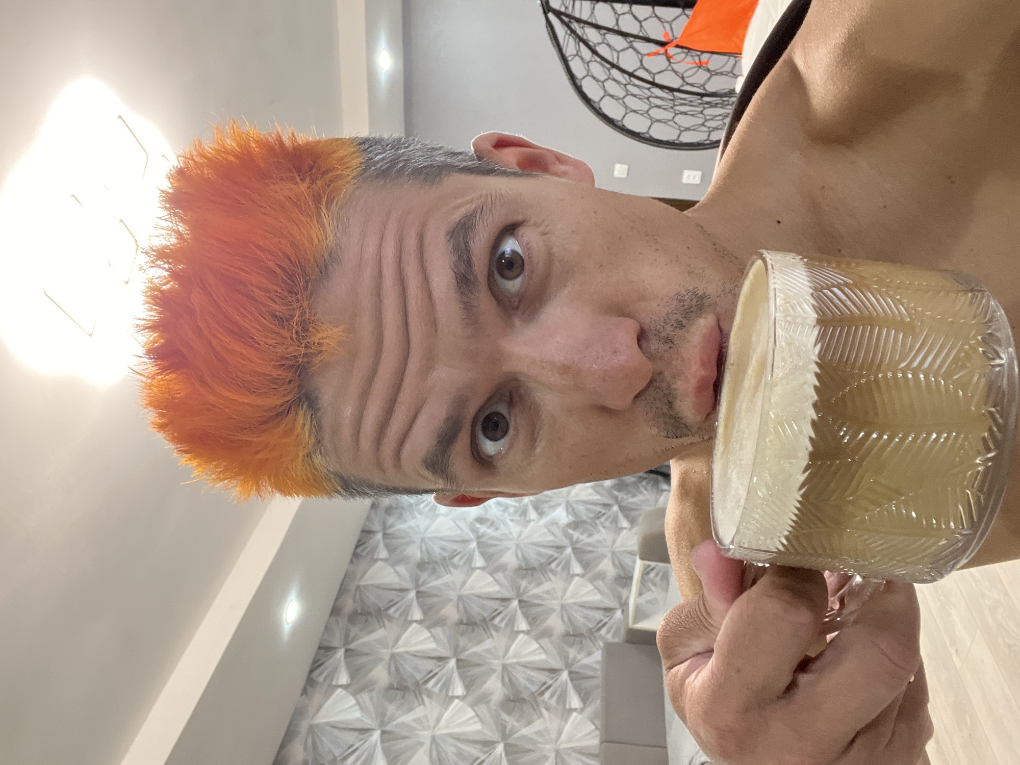 Captured in Chaikovsky, Russia, the image presents a close-up, selfie-style portrait of a man, oriented 90 degrees clockwise from an upright position. The subject is a light-skinned man, appearing middle-aged, with distinctive bright orange hair that transitions into grey at the roots and sides. His wide, brown eyes are fixed directly on the camera, conveying an expression of surprise or intense engagement, complemented by a slightly open mouth and pronounced forehead wrinkles. He has visible stubble on his chin and cheeks and appears to be wearing a dark, possibly sleeveless top.
The man holds a clear glass cup in his right hand. The cup features a distinct textured pattern, possibly depicting palm fronds, and is filled with a light, creamy beverage, likely coffee with milk or a similar drink, topped with foam.
The background indicates a modern indoor setting. To the upper right, a section of a black, intricate hanging chair or basket swing is visible. Behind the man's head, a wall is adorned with a contemporary geometric pattern, suggesting decorative panels or textured wallpaper. The ceiling is white with visible recessed lighting, and a light-colored floor, possibly wood or tile, is seen towards the bottom of the frame. The scene is brightly lit, primarily by artificial light sources. There is no visible text in the image.