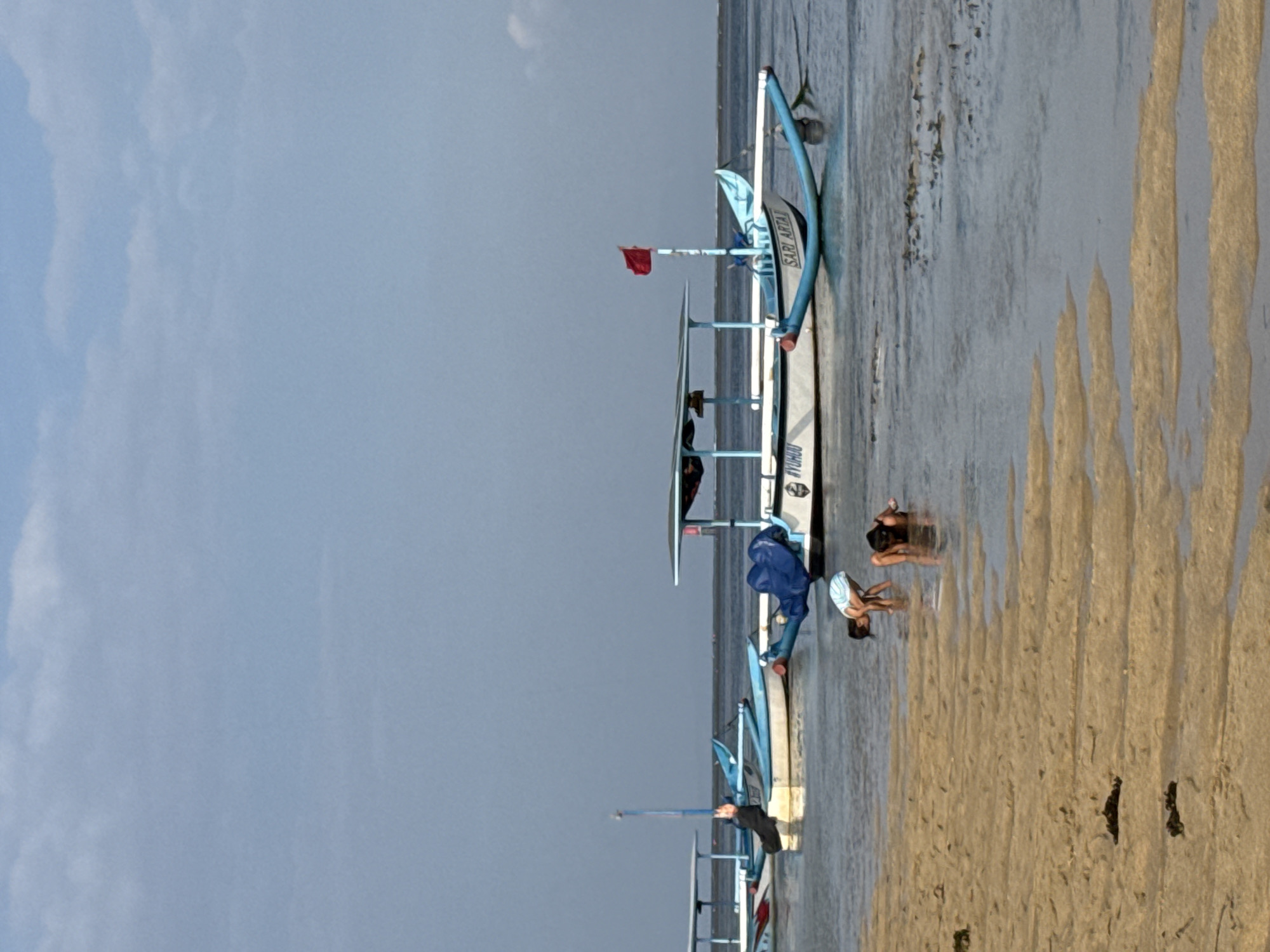 A wide, low-angle shot shows a beach scene with children playing in the shallow water and traditional fishing boats anchored offshore. The sand in the foreground has ripples from the receding tide. In the midground, two children, one wearing a striped top, are wading in the water, bending down as if searching for something. Behind them, several traditional outrigger fishing boats are moored. One boat, closer to the foreground, has a blue canopy, a red flag, and the name "SARI ARTA" painted on its side. Another boat further back is partially covered with a blue tarp. The sky above is a pale blue with scattered clouds. The scene appears to be at Sanur Beach, Denpasar, Indonesia, known for its calm waters and traditional fishing vessels.