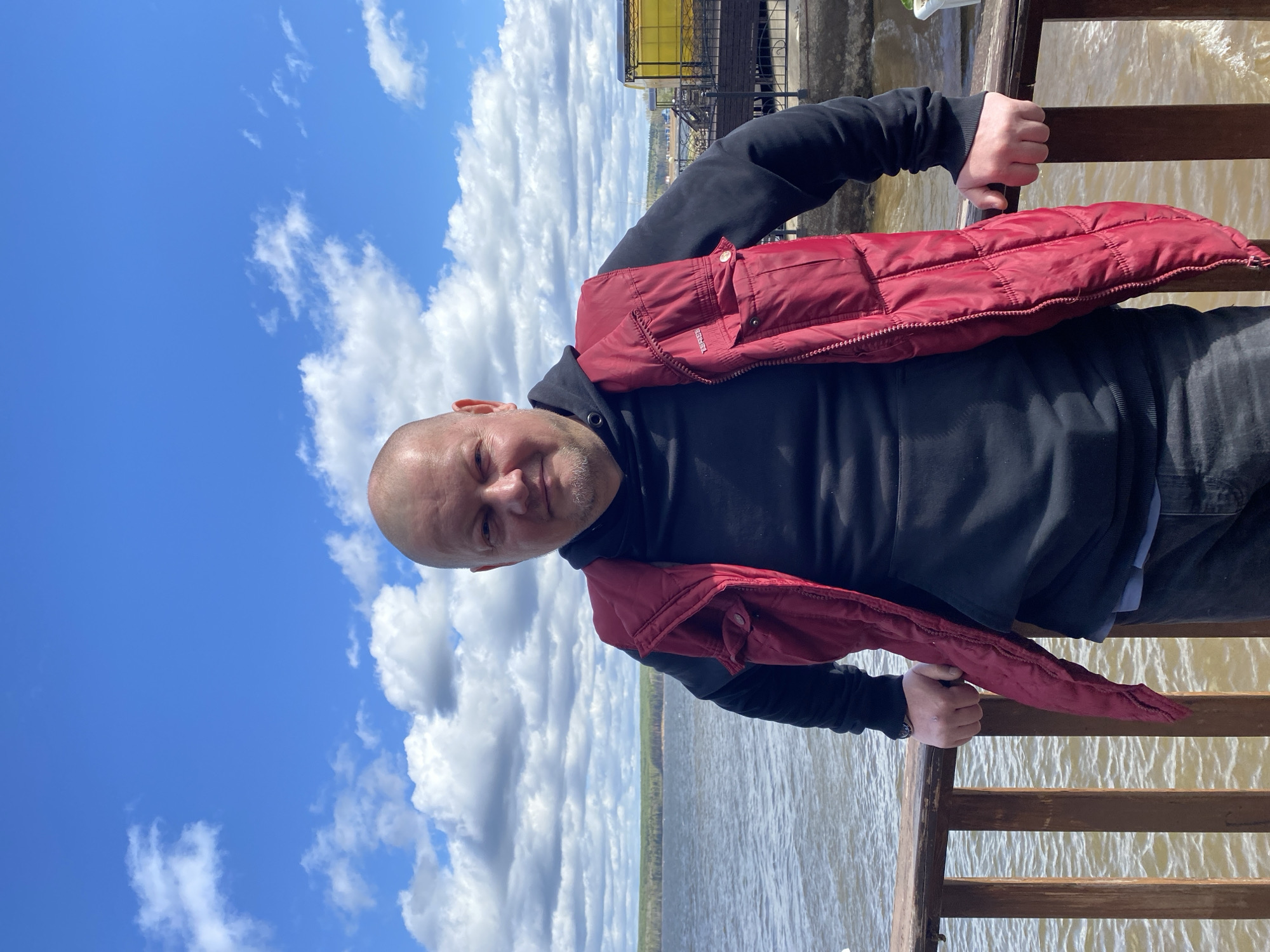 A man stands on a wooden deck or pier in Chaikovsky, Russia, with a body of water and trees in the background. He wears a black hoodie and has a red vest draped over his shoulders. The sky is blue with puffy white clouds. There is a metal structure in the background, potentially part of a building or platform. The man is holding onto the wooden railing of the deck.