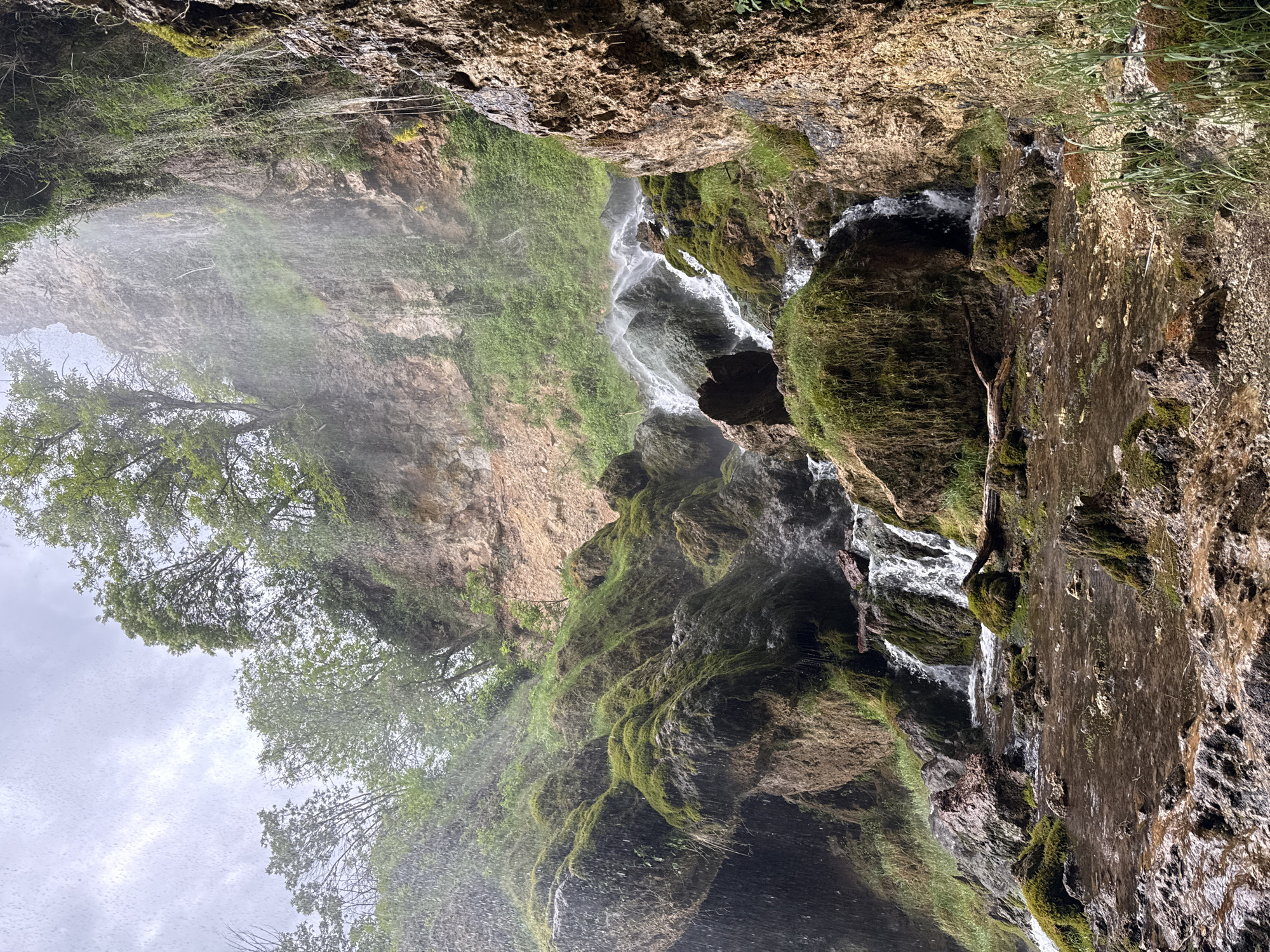 The image depicts a waterfall cascading down a rock face, likely in the vicinity of Razhdavitsa, Bulgaria. The rock face is covered in moss and vegetation. Water streams down the rocks, and spray is visible. Trees are growing near the top of the falls. The sky is overcast.