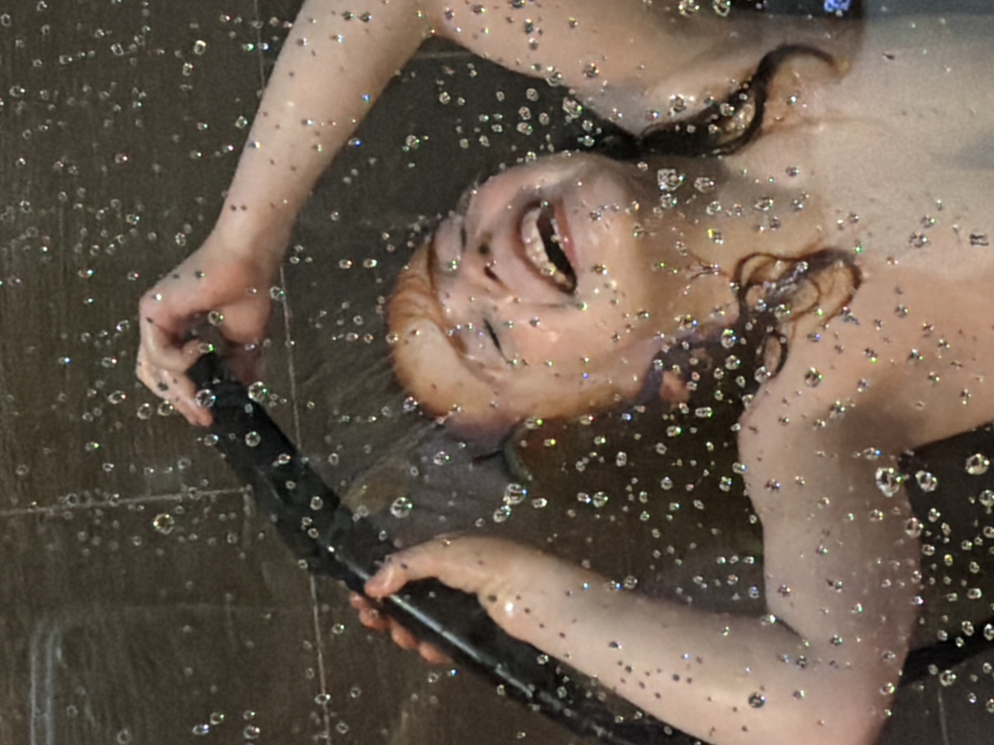 This media file captures a close-up, angled shot of a person in a shower. The person's face is visible, turned upwards and to the left, with their mouth open in what appears to be a laugh or gasp. Their eyes are closed, and their head is tilted back. Water droplets are visible throughout the frame, clinging to the person's skin and hair, and suspended in the air, catching the light. The person's left arm is also visible, holding onto a black shower head. The background appears to be a tiled shower wall. The overall impression is one of playful abandon and enjoyment of the shower. The lighting suggests an indoor setting, likely a bathroom, with the shower running. There is no visible text in the image.